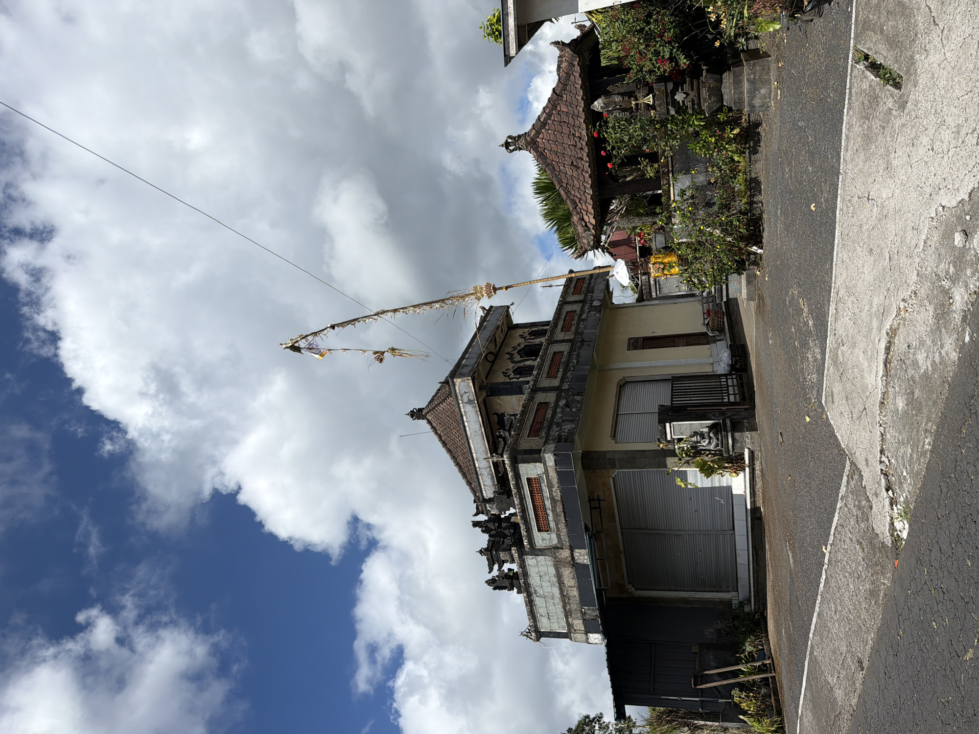 The image captures a street-level view of a building in Kintamani, Indonesia. The building, which appears to be a traditional Balinese structure, features a multi-tiered roof with dark tiles and decorative elements. The facade includes closed roller shutters over what might be shopfronts, and a side entrance with a wooden door. Above the shutters, there are ornate architectural details, including carved figures and patterned vents. To the right of the main building, a smaller, open-air structure with a tiled roof and stone carvings is visible, surrounded by lush greenery and flowering plants. A tall, bamboo-like pole adorned with festive decorations, possibly a Penjor, stands prominently in front of the main building, reaching towards the cloudy blue sky. A power line stretches diagonally across the frame from the upper right. The ground in front of the structures is paved with asphalt and concrete, showing signs of wear. The overall scene suggests a commercial or cultural space in a tropical environment.