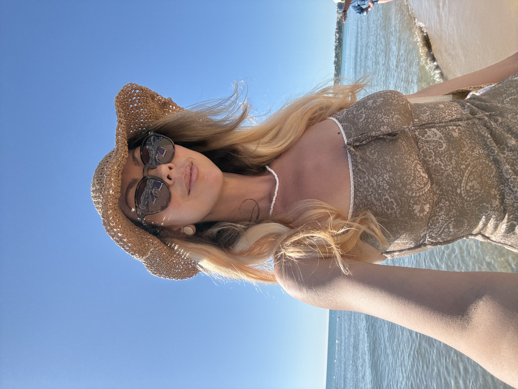 A woman with blonde hair, wearing a straw hat, sunglasses, and a patterned dress, takes a selfie on a beach. The ocean is visible behind her, with a clear blue sky above. In the background, there is a glimpse of a town on a hill. The scene is likely in Nesebar, Bulgaria, given its coastal location and characteristic architecture visible in the distance.