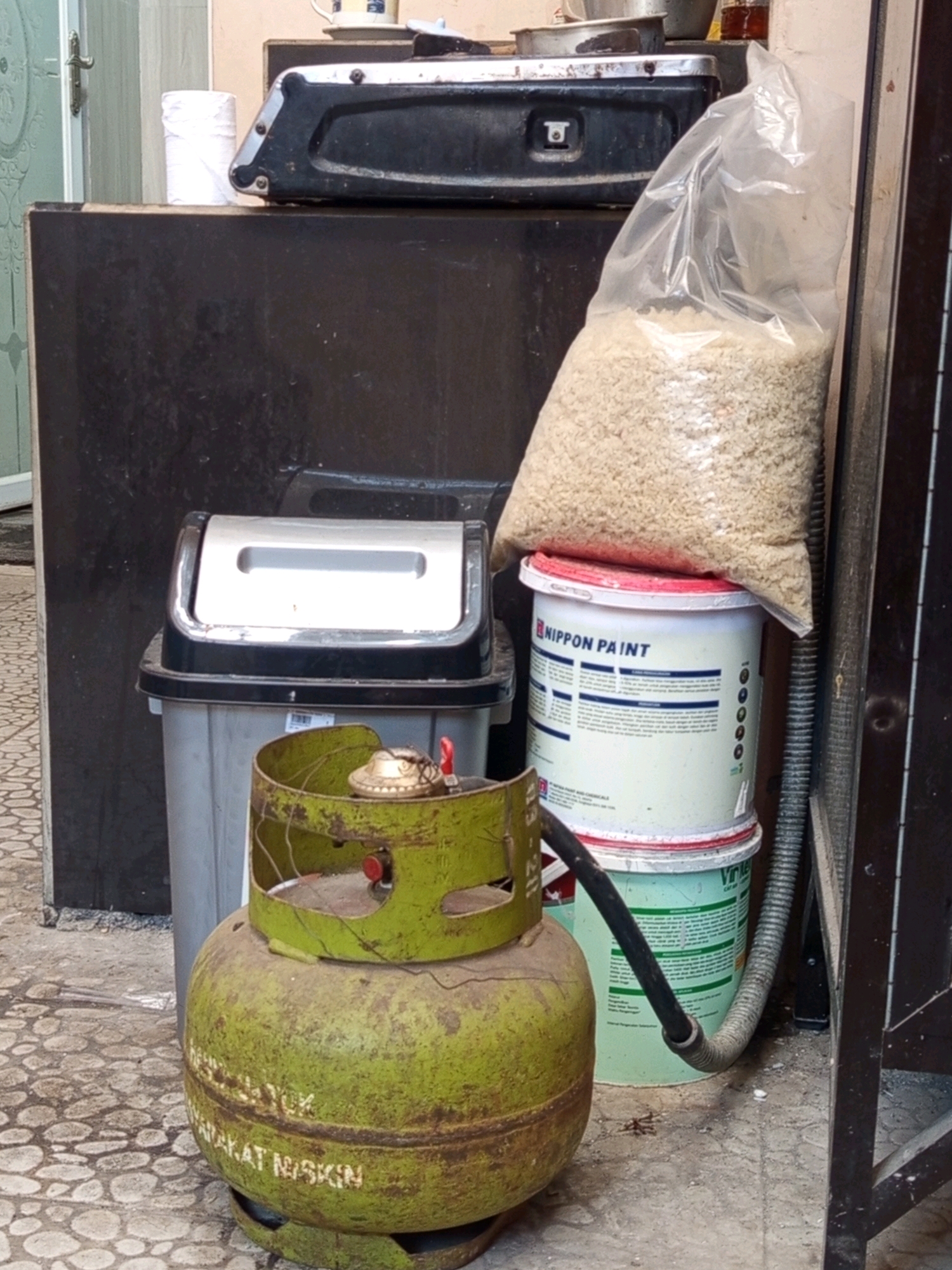 In what appears to be a storage area or alleyway in Malang, Indonesia, a collection of items are visible. In the foreground, a green, round LPG gas cylinder with a yellowed and rusted surface sits on the ground. It has some white text partially visible on its side. Behind the cylinder, a gray trash can with a silver lid is present. To the right of the trash can, two stacked white paint buckets are visible, the top one labeled "NIPPON PAINT". A large, clear plastic bag filled with light-colored granular material, possibly rice, rests on top of the paint buckets and leans against a dark vertical surface. Above the paint buckets and bag of material, a black appliance with a silver edge, resembling a portable stove or grill, is situated on a dark, flat surface. To the left of the stove, a roll of white paper, possibly toilet paper, is positioned on the same dark surface. The ground is paved with light-colored, irregular-shaped stones. A metal fence or gate is partially visible on the far right of the image.