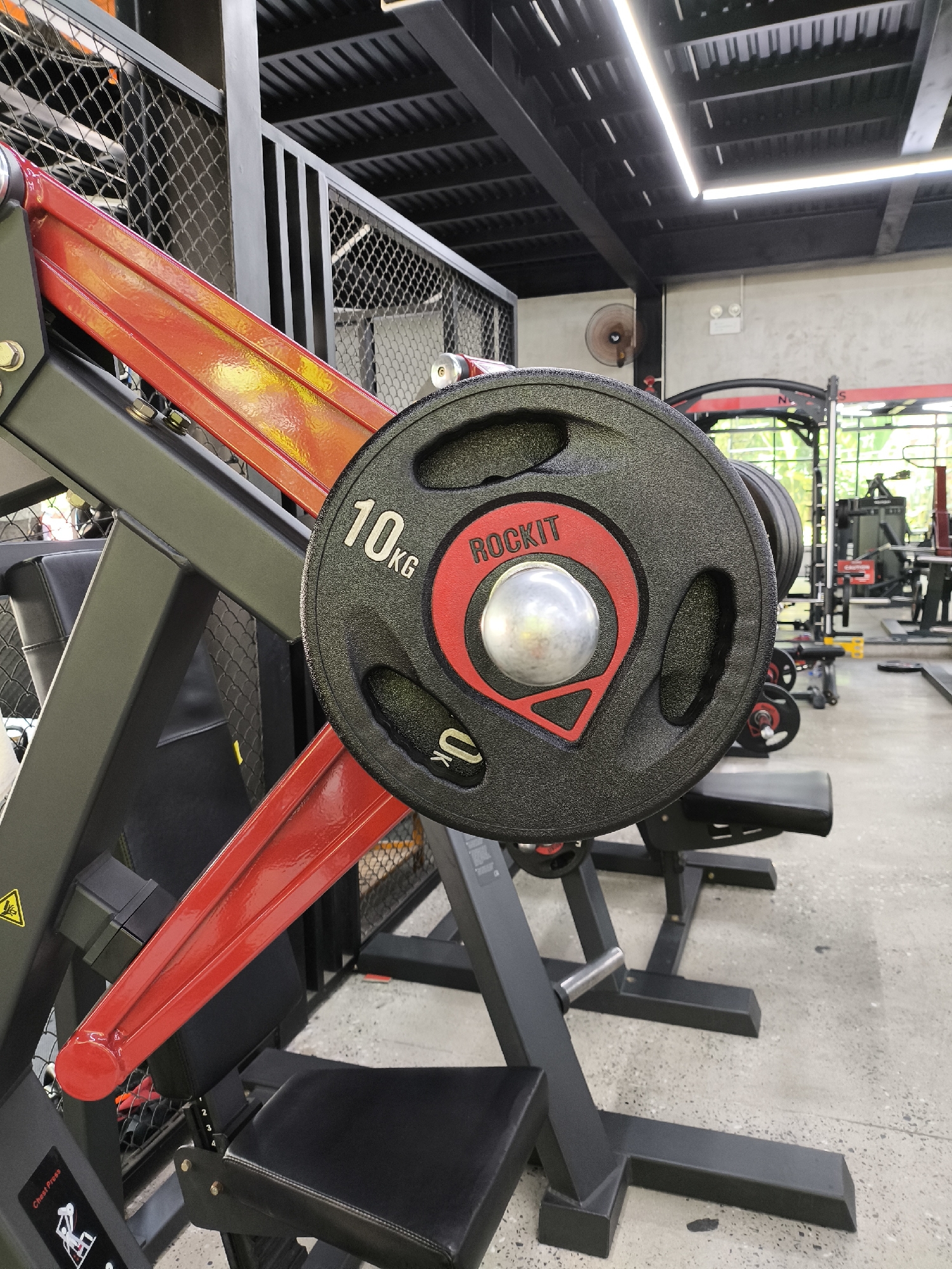 A close-up shot focuses on a black weight plate with red accents and silver lettering that reads "ROCKIT 10KG" on a piece of gym equipment. The equipment itself is black and red, with a padded black seat visible. In the background, other gym equipment, including weight racks and benches, are discernible. The floor is made of concrete. The setting appears to be a gym in Da Nang, Vietnam, given the natural lighting and the style of the gym equipment. There are no people in the frame, and no discernible actions or interactions are taking place. The overall environment is clean and organized, suggesting a well-maintained fitness facility.