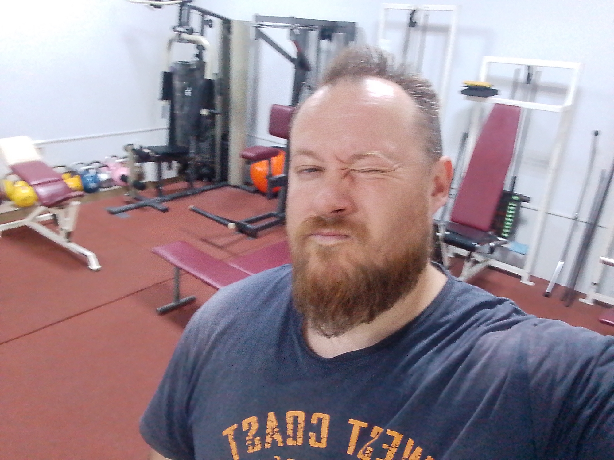 The image shows a man in a gym. He has a beard and is wearing a t-shirt with the words "WEST COAST" on it. He is winking. In the background, there is gym equipment, including a weight machine, a bench, and kettlebells. The floor is red. The location is likely Chaikovsky, Russia.