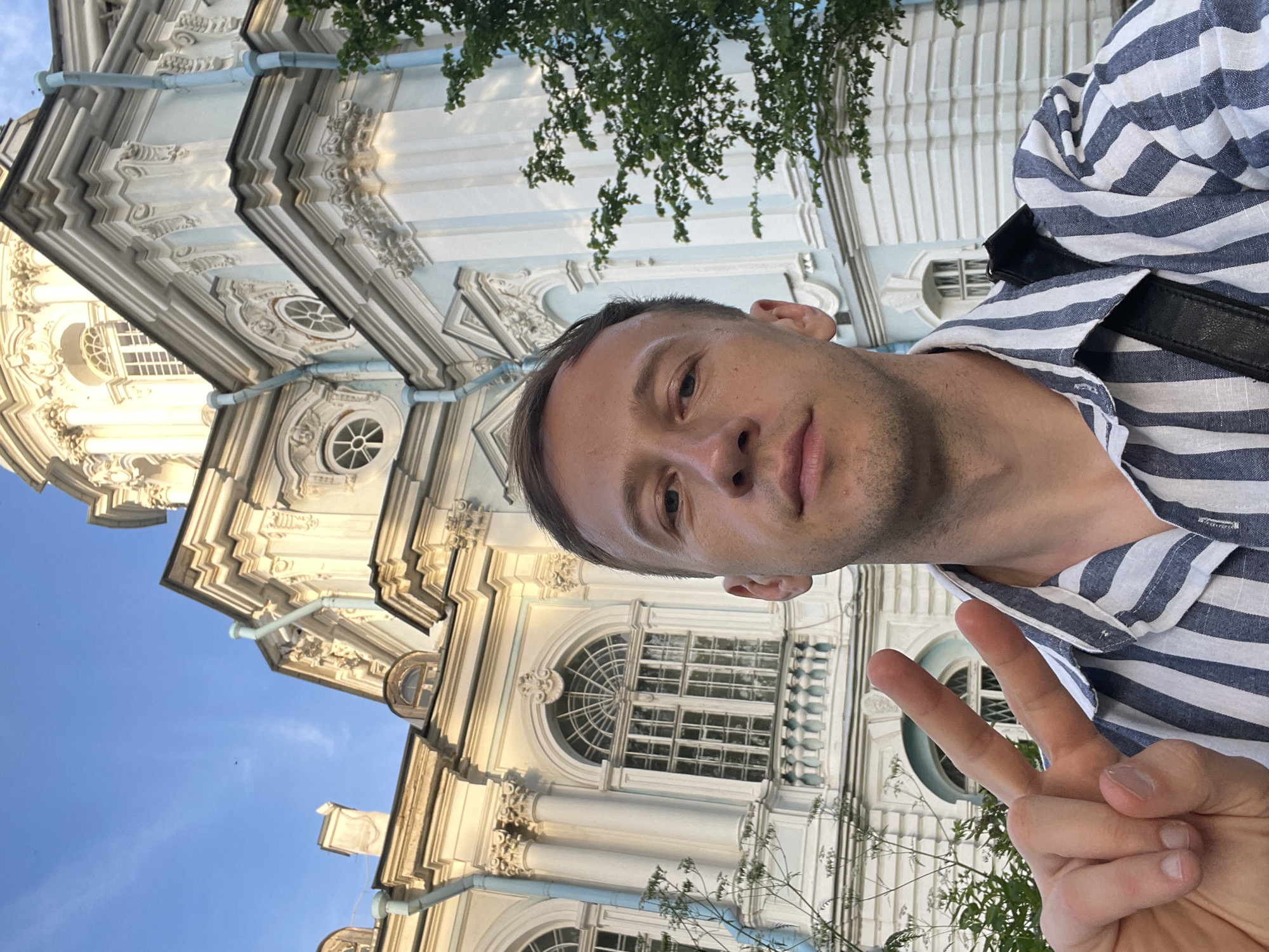 A male subject is captured in a close-up selfie, positioned centrally and looking directly at the camera. He has short brown hair and is wearing a blue and white vertically striped collared shirt, with a black strap from a bag visible over his right shoulder. His left hand is raised, displaying a V-sign with the index and middle fingers. The background features a prominent, ornate Baroque-style building, predominantly off-white or light cream, with light blue architectural accents on columns, window frames, and decorative elements. Visible architectural details include large arched windows with grilles, circular windows, balustrades, decorative cornices, and light blue vertical drainpipes. A large dome is partially visible at the top, suggesting a cathedral or similar structure. The sky above is clear blue with faint wisps of white clouds, indicating daytime. Green foliage from trees and shrubs is present on both sides of the frame. This scene is situated in the city of Unknown, Russia.