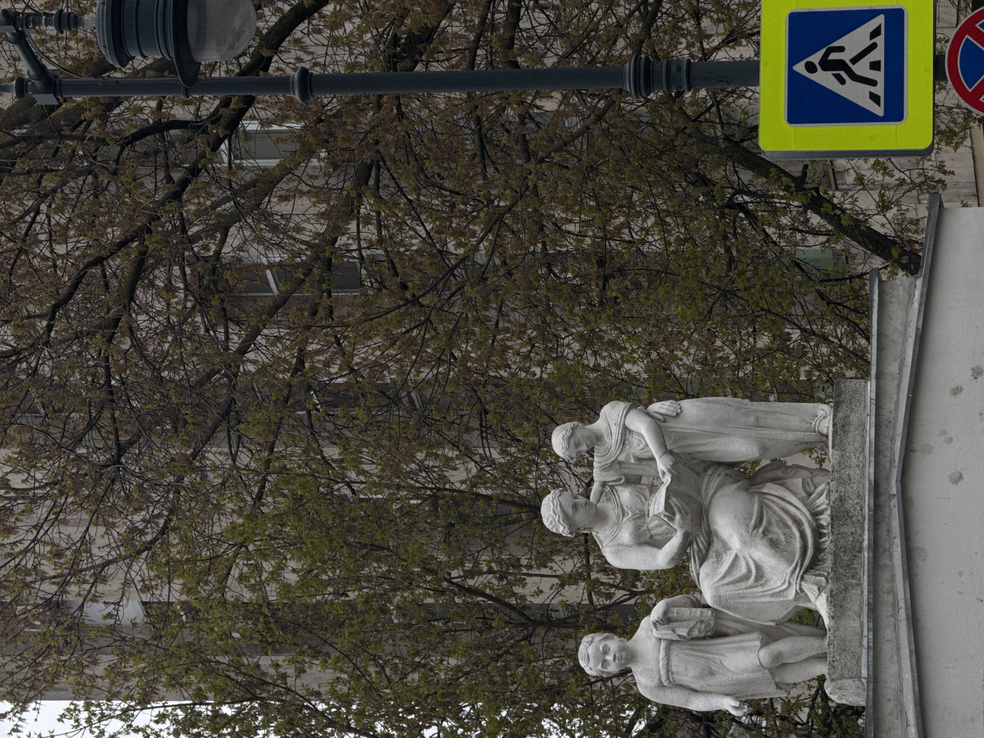 The image depicts a stone sculpture of three figures, likely women, in front of a building partially obscured by a tree with budding leaves. Above the sculpture, there's a pedestrian crossing sign and a street lamp. The sculpture appears to be placed on a plinth or pedestal. Given the architecture and street signs, the location is likely in Moscow, Russia. The statue is in the foreground and partially obscures the background, in which there is an office building.