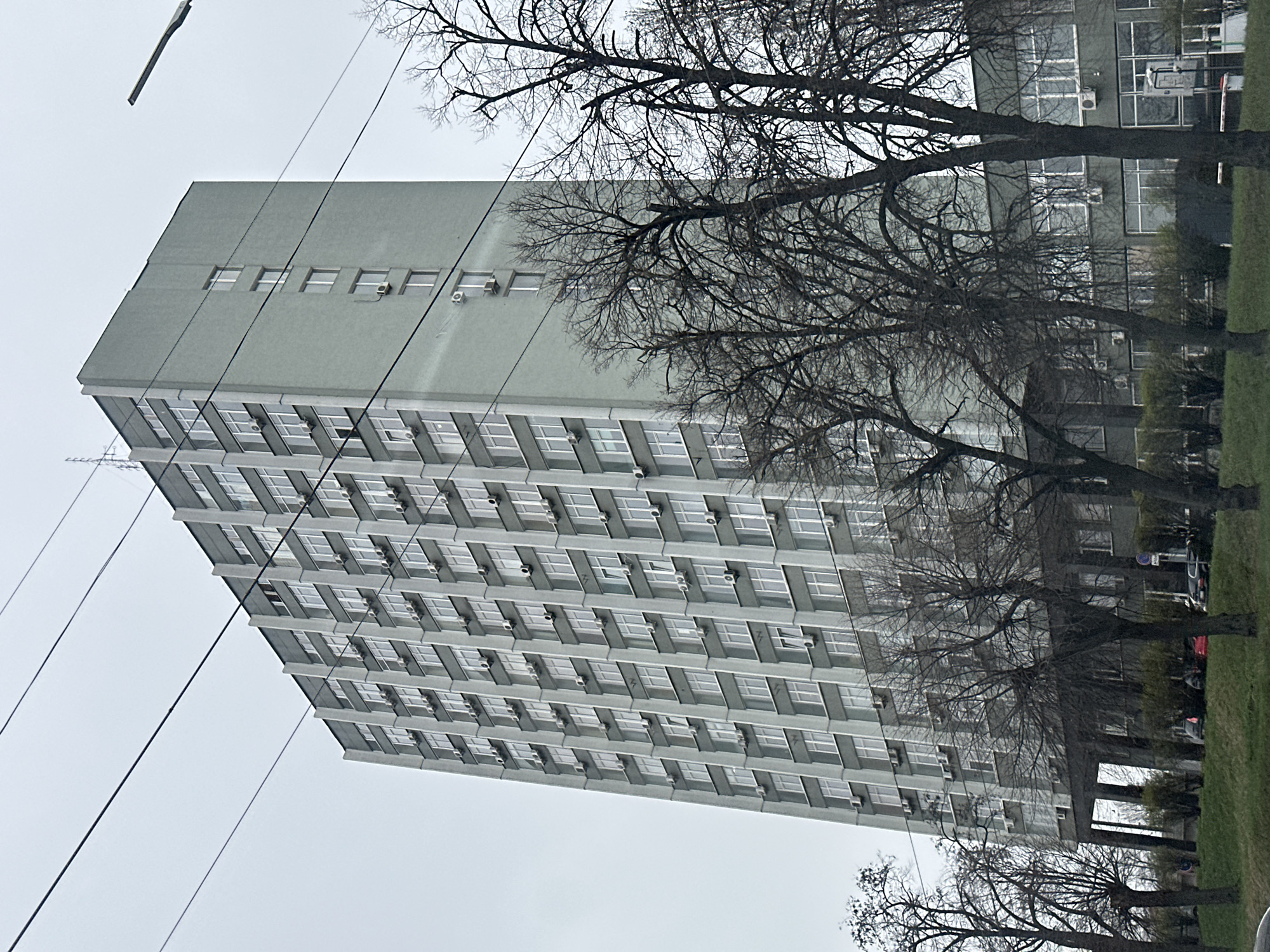 The image captures a multi-story, possibly institutional, building located in Kyiv, Ukraine. The building is predominantly gray, with a repetitive pattern of windows and externally mounted air conditioning units on most floors. It appears to be a concrete structure, possibly dating back to the Soviet era. The sky is overcast and gray, suggesting dull weather. Bare tree branches are in the foreground, partially obscuring the view of the building. Utility wires crisscross the scene. Ground level has some vegetation, and a hint of parked cars suggests a street or parking area. No people are visible in the image. The overall impression is one of urban austerity and bleak weather.