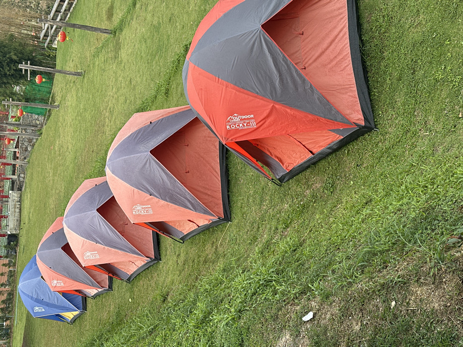 Six dome-style camping tents are pitched in a diagonal line on a green, grassy slope in Ban Mai Saha Sampan, Thailand. The leftmost tent is blue with yellow accents. The subsequent five tents are orange and dark gray, with "OUTDOOR ROCKY-II" branding visible on several. No individuals are present. The tents are arranged on a well-maintained lawn that slopes upward towards the background. In the background, beyond the tents, there are structures with red roofs and railings, resembling traditional architecture. Multiple red spherical lanterns hang from wooden posts and structures in the mid-ground and background. Additional green foliage and wooden fencing are visible on the right side of the frame. The ground immediately adjacent to the tents on the left exhibits wilder grass and some exposed soil.