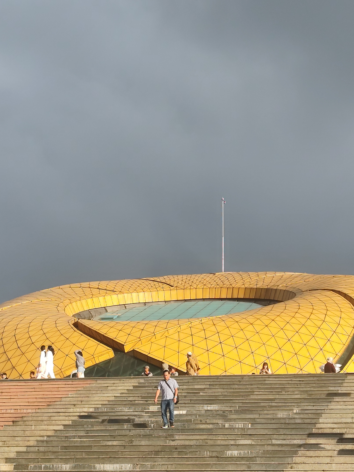 This image depicts the exterior of the Da Lat Exhibition Center, a striking modern building with a distinctive golden, honeycomb-patterned roof. The structure features a large, circular opening in the roof, revealing a pool of water or a reflective surface. The building is situated in Da Lat, Vietnam, a city known for its colonial architecture and natural beauty.
In the foreground, a wide set of concrete stairs leads up towards the building. Several people are visible on the stairs and on the elevated plaza in front of the building. A man in a light-colored shirt and jeans is walking down the stairs. Further up the stairs, a group of people, including two women in white, are walking together. Other individuals are scattered across the plaza and stairs, some standing and some sitting.
The sky above is overcast and a muted gray, suggesting a cloudy or possibly approaching storm. A flagpole with a flag is visible in the distance, rising above the golden roof. The overall scene captures a moment of everyday life against the backdrop of a prominent architectural landmark.