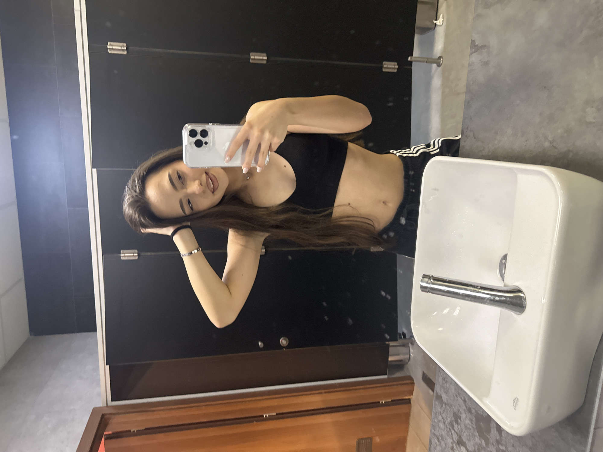In an indoor public restroom with black stall doors and a white sink, a young woman with long brown hair takes a selfie in a mirror. She is wearing a black sports bra and black athletic shorts with white stripes. She is holding her phone in her right hand, angled to capture her reflection. Her left hand is raised to her head, touching her hair. The sink is white and rectangular, with a chrome faucet. The walls are tiled in gray and black. The lighting is artificial, casting a soft glow on the scene.