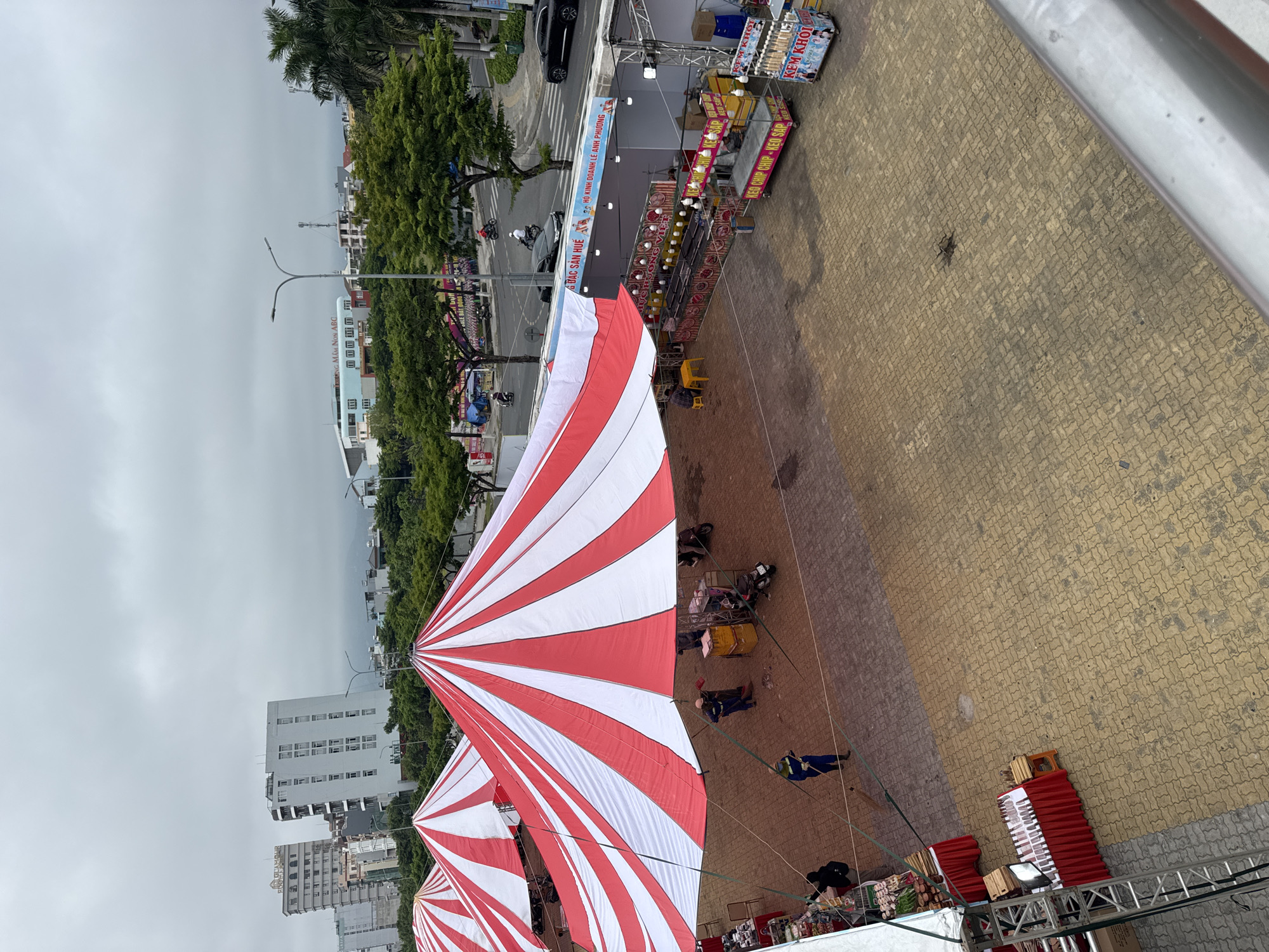 The image, taken in Đà Nẵng, Vietnam, presents a high-angle view of a bustling street fair or market under red and white striped tents. Vendors are seen setting up stalls, and people are walking around or interacting with the stalls. The foreground includes a partial view of the structure from which the photo was taken, likely a balcony or high platform. The marketplace features diverse stalls selling food and drinks, indicated by signs reading "KEM KHOI" and "KEO CHIP CHIP - KEO SAP" ("Ice Cream" and "Chip Chip Candy - Sticky Candy"), as well as a sign that translates to "Specialty Products of Hue," indicating the presence of regional Vietnamese goods. Further into the scene, a street with light traffic is visible, alongside trees and buildings under an overcast sky. A sign for "MAM NON ABC" (ABC Kindergarten) can be seen among the buildings. The overall setting portrays a busy public space during market preparation.