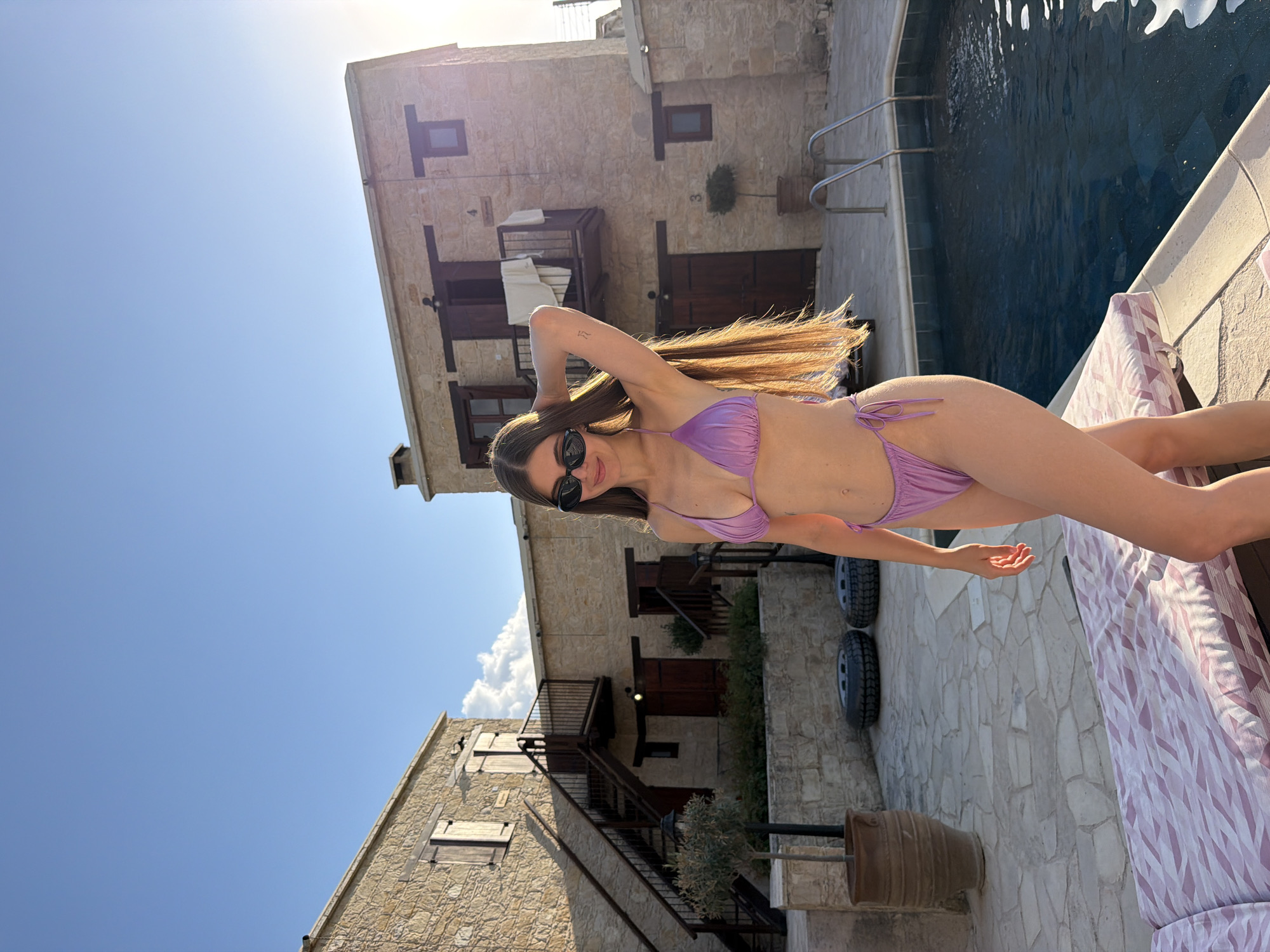 A young woman with long, brown hair and sunglasses is posing by a swimming pool. She is wearing a purple bikini and is leaning back with one hand behind her head. The setting appears to be a courtyard with stone buildings, possibly an old complex or hotel, with balconies and wooden shutters. A swimming pool is to her right, with metal railings leading into the water. The sun is bright, suggesting it is daytime, and the sky is a clear blue. The overall mood seems relaxed and summery. There are also some decorative elements like a large terracotta pot and two tires visible in the scene. The woman's pose and smile suggest a sense of leisure and enjoyment. The buildings have a rustic, traditional architectural style.