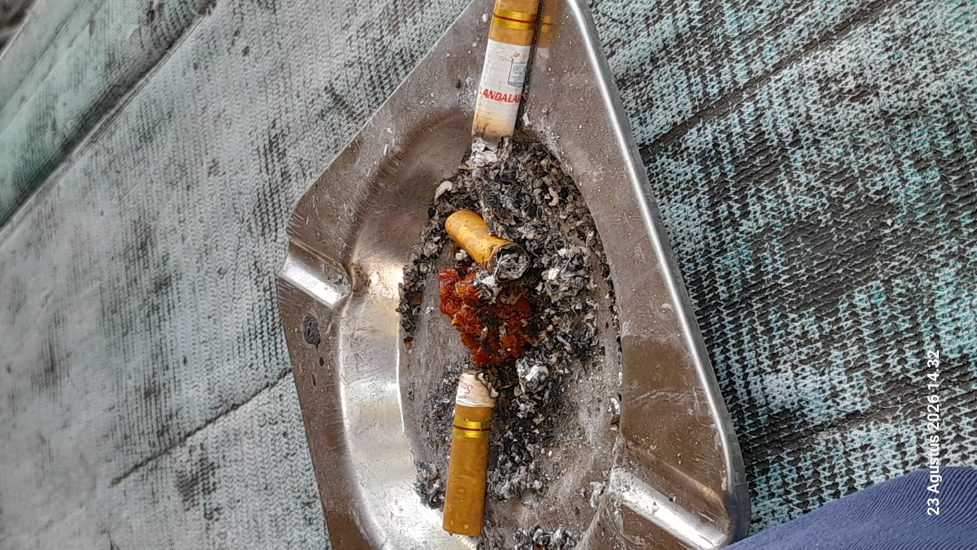 An ashtray containing cigarette butts and ash is placed on a textured surface. There are two cigarette butts in the ashtray, one with a red and gold band and text that appears to read "ANDALAN". The other cigarette butt is shorter and appears to have been recently extinguished. In the center of the ashtray is a small pile of what looks like dried red berries or seeds. The background surface has a woven texture with alternating dark and light blue threads. The lower left corner of the image displays the date and time: "23 Agustus 2026 14.32".