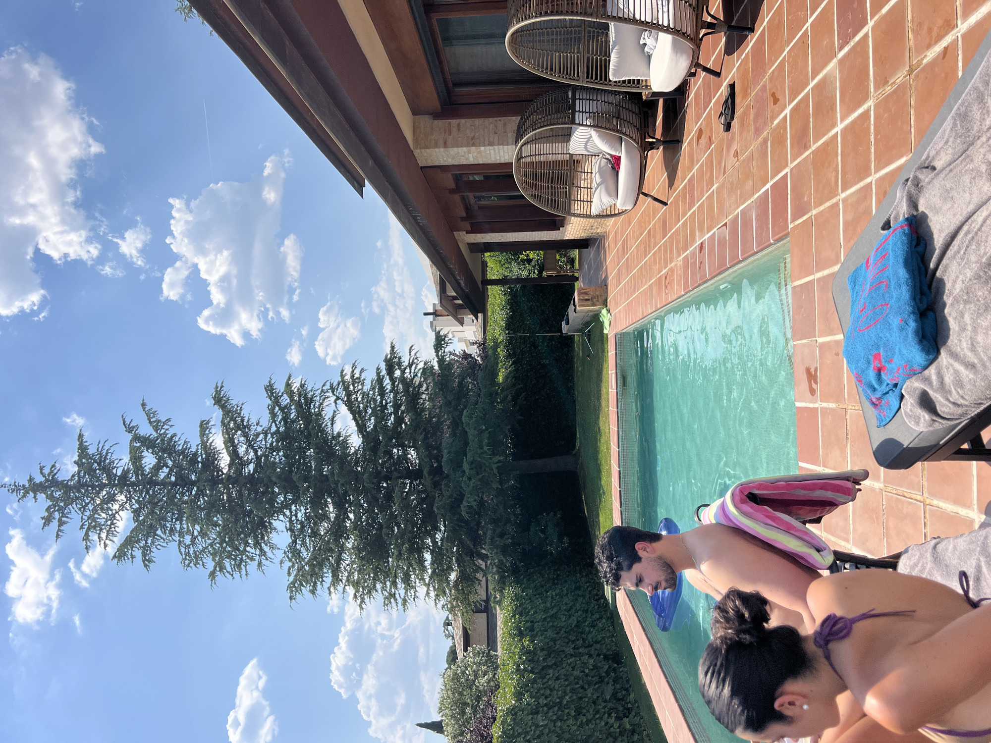 A medium-high angle shot depicts a sunny scene by a swimming pool. Two individuals are partially visible in the foreground near the pool's edge; one is a man with dark hair, and the other is a woman in a purple bikini. They are both looking down, possibly into the pool. A blue towel and a striped towel are placed on a nearby lounger. The swimming pool's water is a clear, light blue-green. Along the side of the pool is a well-maintained hedge. Beyond the hedge, a large tree dominates the landscape, with a clear blue sky dotted with white clouds visible behind it. To the right of the pool, the patio is tiled in terracotta. Two rattan chairs with white cushions are positioned on the patio. The side of a house with brown accents and a covered patio is visible above the patio and chairs. The location appears to be a private backyard, potentially in a residential area outside the city center. Without additional evidence, it is difficult to confirm the exact location in Madrid, Spain, but the architecture and vegetation suggest a suburban or rural area around Madrid.