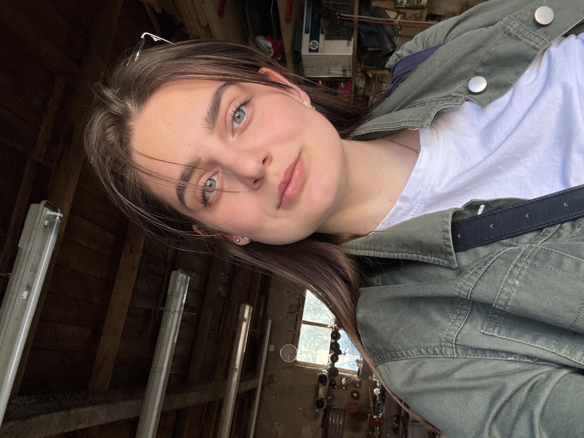 A woman is taking a selfie indoors. She has fair skin, light eyes, and brown hair, wearing a white t-shirt and a green button-down jacket. The background appears to be an unfinished attic or storage space, with exposed wooden beams, fluorescent light fixtures, and various stored items. The location, Trnava, Slovakia, is not evident from the provided image.