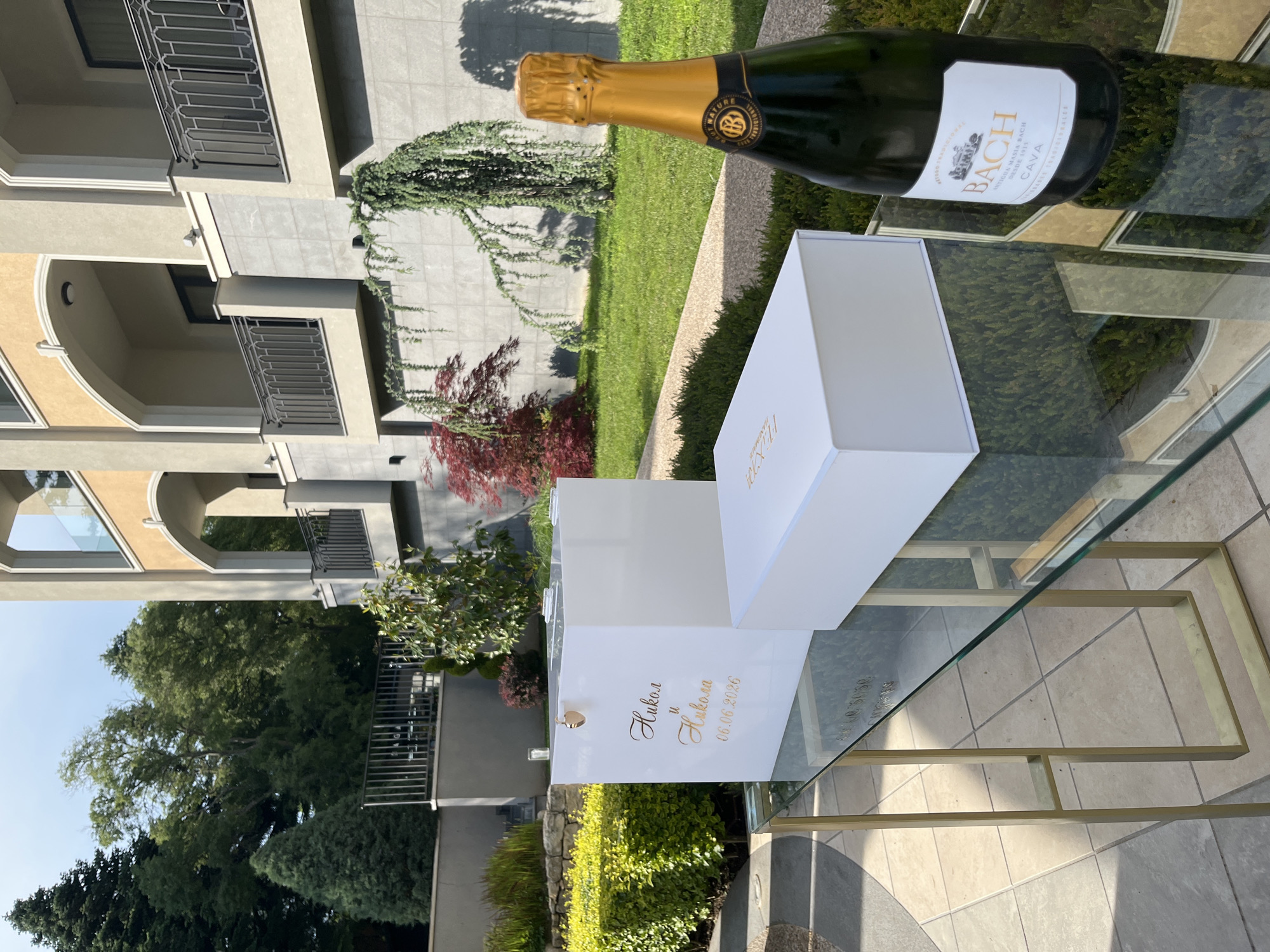The image depicts an outdoor celebratory setup, likely a wedding, in a well-landscaped environment. The scene is illuminated by bright natural daylight, casting short shadows and suggesting a midday to early afternoon time. While not explicitly visible in the image, the location is specified as Varna, Bulgaria.
In the foreground, a modern glass-top table with a golden-colored geometric metal frame stands on light-colored square paving stones. On the table, from left to right, are:
1. A tall, rectangular white box with a clear top opening, featuring a small golden heart-shaped clasp or lock. The front of this box is adorned with gold script text that reads "Никол и Никола" (Nikol and Nikola) followed by the date "06.06.2020". This box appears to be a card or gift reception box.
2. A shorter, rectangular white gift box, positioned to the right of the taller box. This box also has faint gold branding on its top surface, partially visible, and appears to be branded "BACH" on the side.
3. A dark green bottle of sparkling wine, specifically Cava, positioned to the right. The gold foil neck covering is intact. The label clearly identifies the brand as "BACH", type as "BRUT NATURE", and includes "ANTIGUA MASIA BACH DESDE 1915".
The midground features a meticulously maintained garden area. Immediately behind the table, a low, dense green hedge is visible. Beyond this, there is a stone retaining wall and further landscaping, including manicured shrubs, a small tree with reddish leaves, and a trailing green plant (possibly a weeping juniper) growing against a grey tiled wall. A well-maintained green lawn extends into the background.
The background is dominated by a multi-story building, likely a modern residential complex or hotel. The architecture features a combination of yellow/beige upper sections with arched balconies and windows, and grey tiled lower sections. The balconies are equipped with dark metal railings exhibiting decorative patterns. Tall, mature green trees are visible behind and to the left of the building, under a clear blue sky. No individuals are present in the scene.