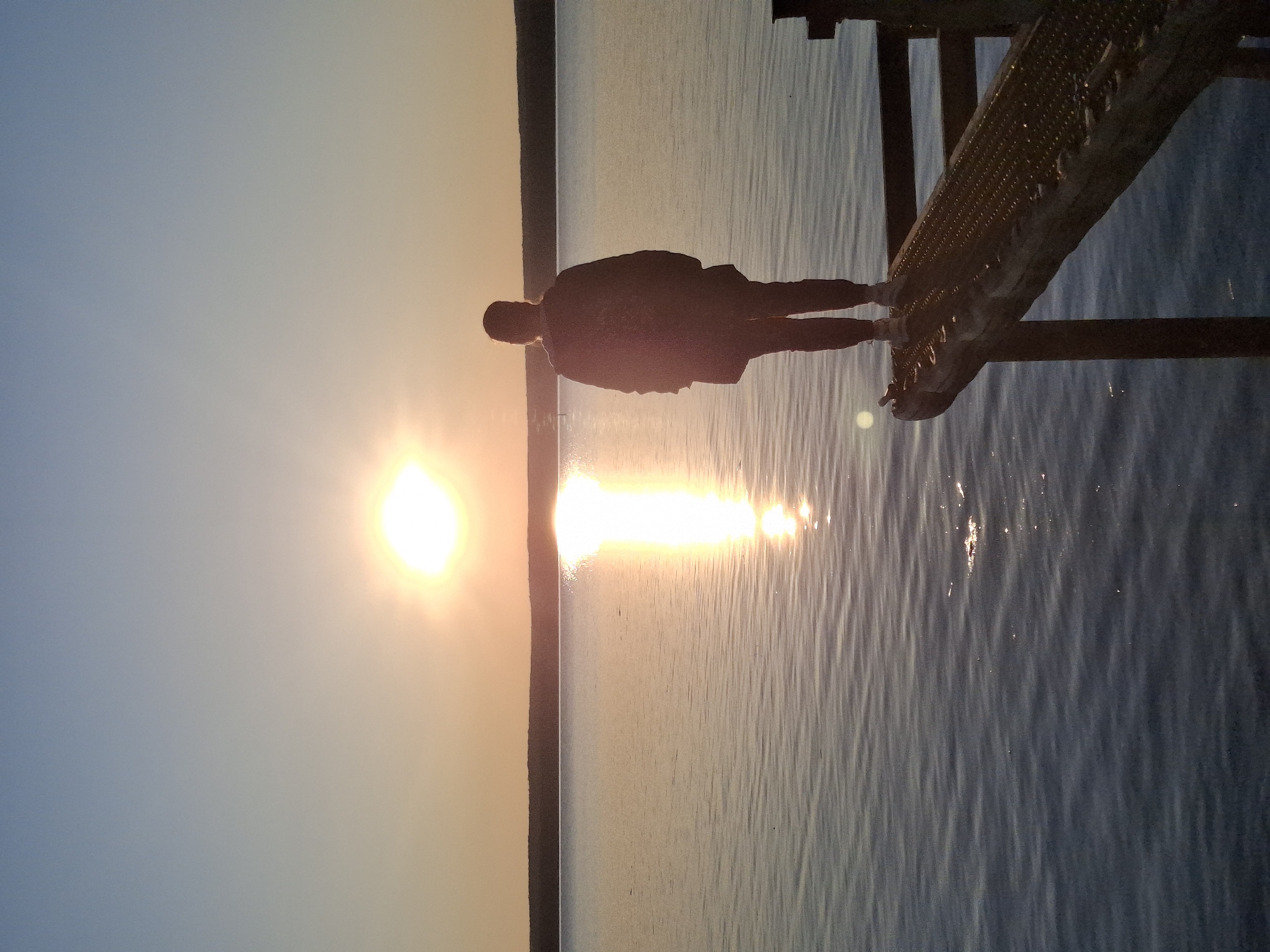 The image depicts a silhouetted individual standing on a metal structure, likely a pier or dock, over a body of water. The sun is setting, creating a bright reflection on the water's surface. The location is the city of Chaikovsky, Russia. The sky is a gradient of blue to orange/yellow near the horizon. The water is calm with ripples and sunlit reflections. The metal structure appears to be made of a perforated material supported by beams.