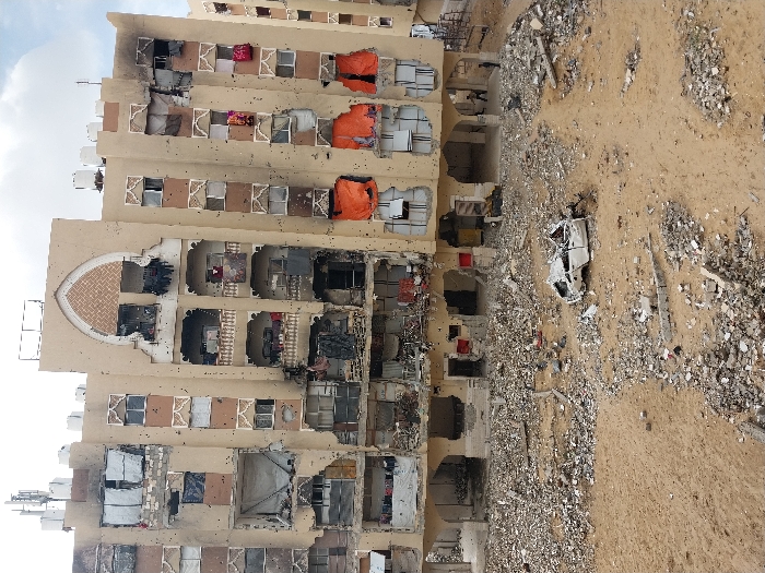 This image depicts a severely damaged multi-story building in Khan Yunis, Palestinian Territory, under an overcast sky. The building, constructed with a light brown facade and featuring an arched decorative motif centrally on its upper stories, exhibits extensive structural damage. Numerous windows are blown out or boarded up with various materials, including corrugated metal and fabric. The facade is pockmarked with numerous small, circular impact marks, consistent with projectile strikes. Several sections show charring, particularly on the lower-mid levels and some balcony areas, indicating fire damage. Balconies contain debris and hanging remnants of clothing. The ground immediately in front of the building is a desolate expanse of sand and loose dirt, heavily strewn with large quantities of building rubble, concrete fragments, and twisted metal debris. A completely destroyed white vehicle, reduced to a crumpled chassis and interior components, lies centrally on the debris-strewn ground. In the distance, near the building's ground floor entrance, two individuals are visible; one stands upright, while the other appears to be in motion or partially obscured.