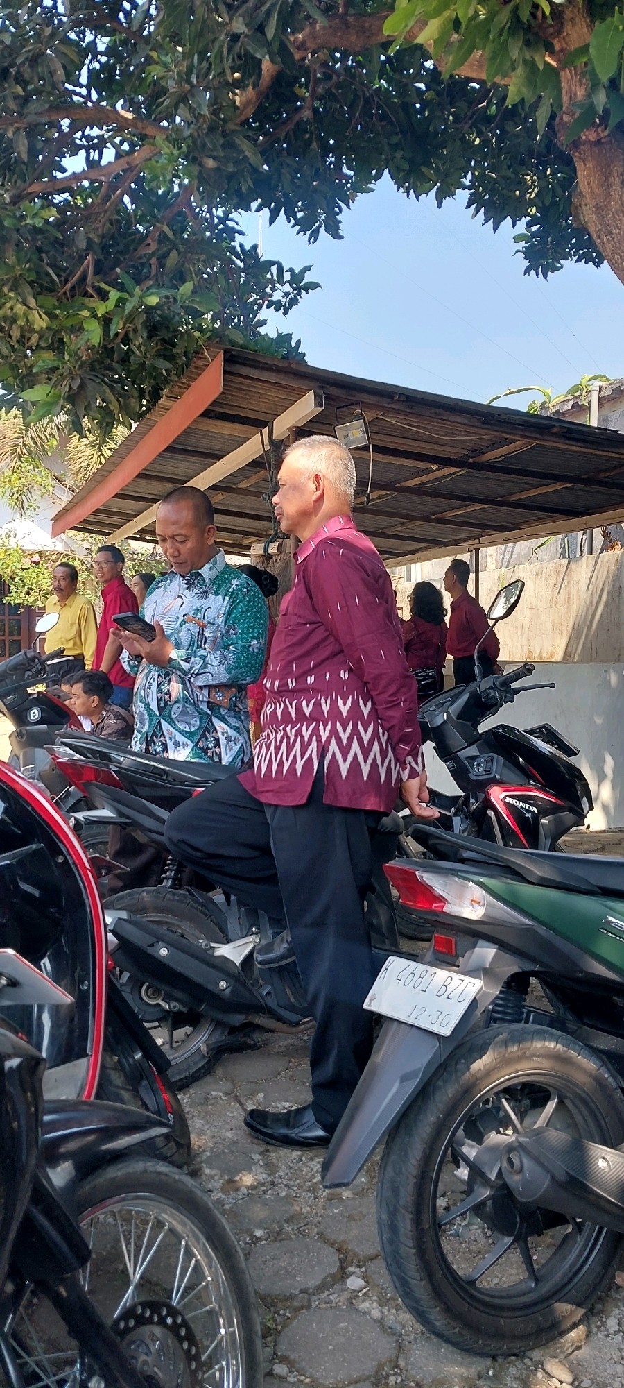 In Keling, Indonesia, under the dappled shade of a tree, a group of men gather near a row of parked motorcycles. A man in a teal and white patterned shirt holds a smartphone and appears to be showing it to another man dressed in a burgundy shirt with a geometric pattern. The man in burgundy stands next to a black motorcycle, his hands clasped in front of him. Behind them, other individuals are visible, some in red shirts and one in a yellow shirt. The environment appears to be an outdoor gathering, possibly at a public space or an event, with a simple corrugated metal roof structure providing additional shade. The ground is paved with interlocking grey stones.