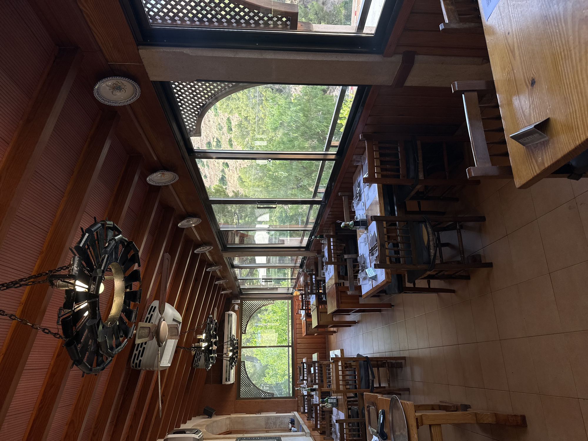 This image depicts the interior of a restaurant in Gènova, Spain. The restaurant features wooden tables and chairs, with many tables set for dining. Large windows line the walls, offering a view of lush greenery and a hillside. Decorative plates are mounted on the wooden ceiling beams, and a unique circular chandelier hangs above the dining area. Air conditioning units are visible on the ceiling as well. The atmosphere appears to be calm and ready for patrons.