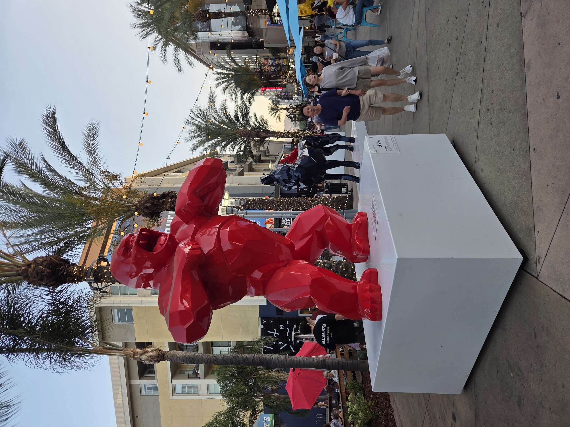 The image depicts an outdoor scene at dusk, likely in a commercial or entertainment district. A large, faceted red sculpture of a gorilla is prominently displayed on a white pedestal in the foreground. To the left of the gorilla, several people are seated at tables outside a venue, with red umbrellas providing shade. Palm trees with decorative lights are interspersed throughout the scene, and string lights are hung across the street, illuminating the area.
In the middle ground, a black sculpture of a horse stands near the gorilla. A couple walks hand-in-hand past the sculptures, with other pedestrians visible in the background. Buildings line both sides of the street, featuring storefronts and balconies. The overall atmosphere is lively and suggests a public gathering space. The location is identified as Burbank, United States of America.