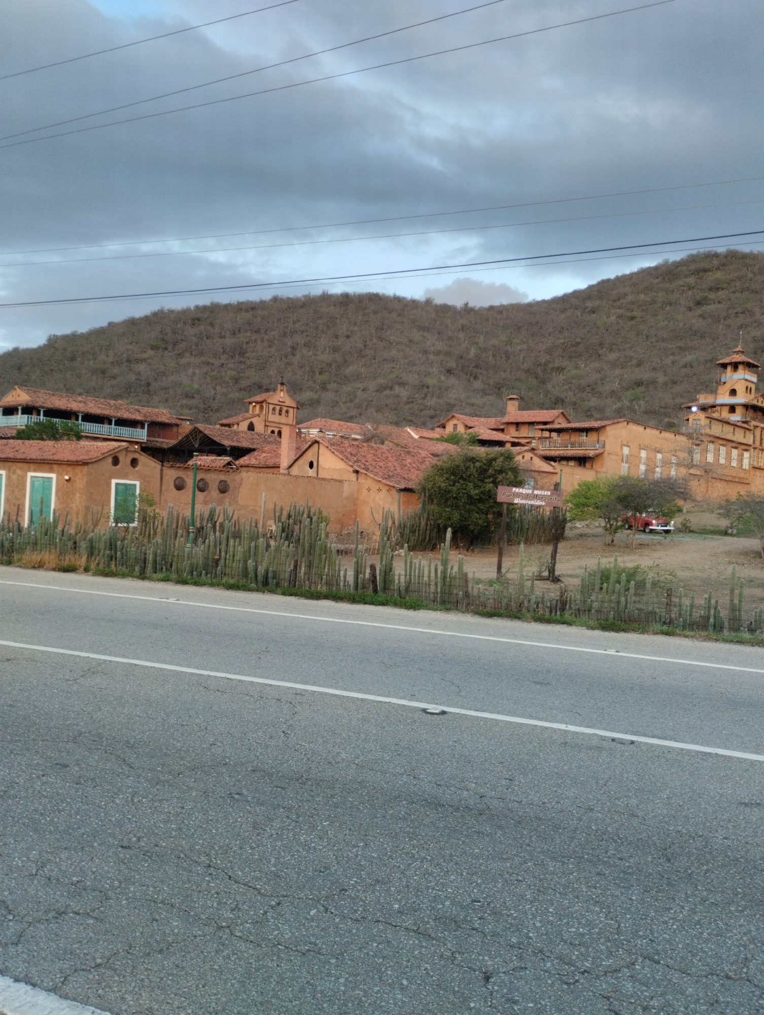 The image depicts a view of the "Parque Museo Pueblos de Margarita" in Juan Griego, Venezuela. In the foreground, a cracked asphalt road with a white lane marker runs horizontally across the frame. Behind the road, a line of tall, slender cacti stands in front of a fence. Beyond the cacti, several ochre-colored buildings with red tile roofs are visible. These buildings appear to be part of a historical or cultural complex, with architectural details like balconies, arched windows, and a tower resembling a bell tower. The structures are situated at the base of a barren, brown hillside covered in dry brush. The sky above is overcast with dark, heavy clouds. Power lines are strung across the upper portion of the image. A red vehicle is parked in the dirt lot behind the buildings, and a wooden sign, partially obscured, indicates "PARQUE MUSEO PUEBLOS de MARGARITA Bienvenidos." The overall scene conveys a sense of arid, historical landscape.