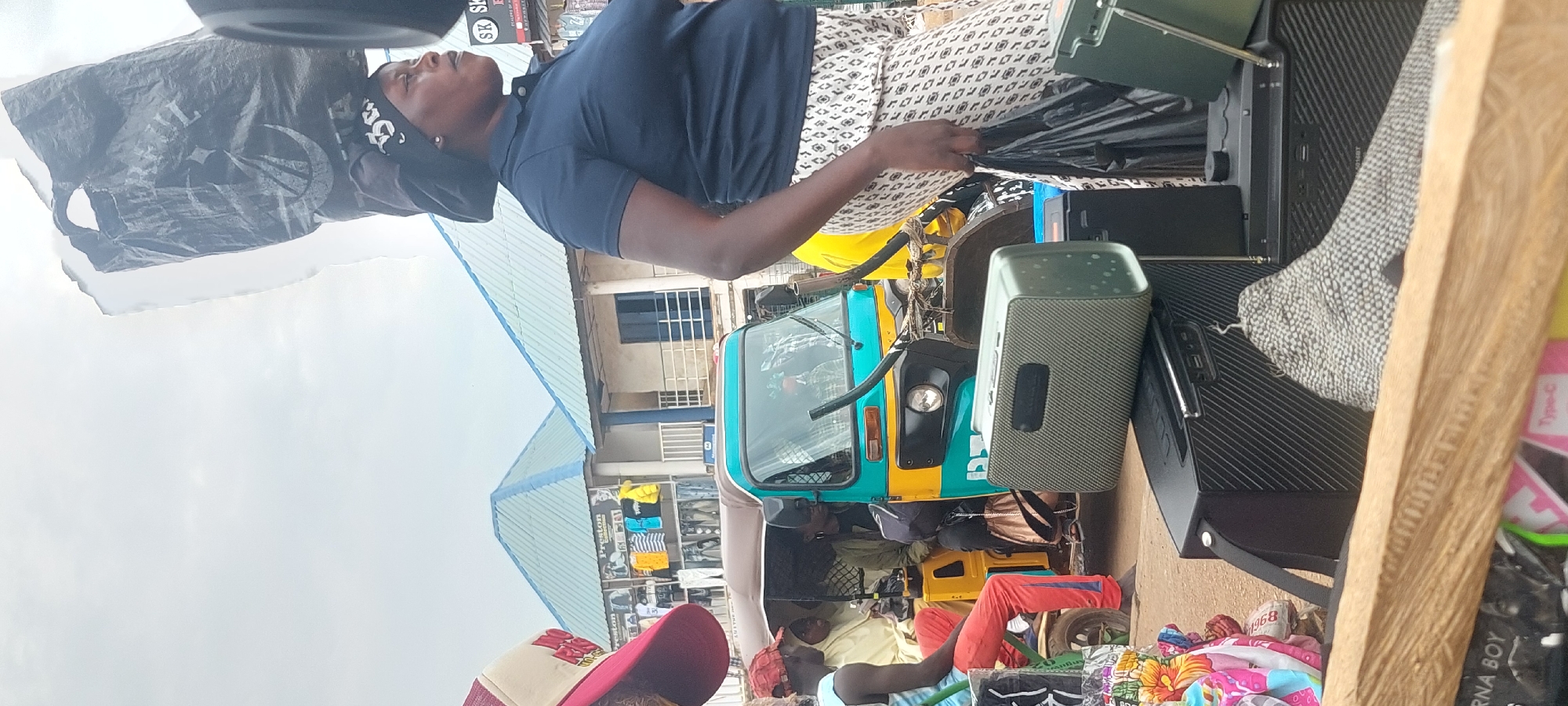An outdoor market scene features a woman carrying a large black plastic bag on her head, wearing a dark blue polo shirt and a patterned skirt. She holds another black plastic bag in her right hand. In the foreground, two speakers are visible, one light green and ribbed, the other black and ribbed. Behind the woman, a turquoise and yellow three-wheeled vehicle, possibly a tuk-tuk, is partially obscured. Several people are seated inside the vehicle. The background shows buildings with light blue roofs and a stall displaying clothing. The ground is dusty or dirt. The sky is overcast.