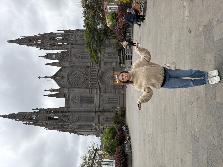 A young Caucasian female stands centrally in a large, paved public square, identified as the plaza in front of the Church of San Juan Bautista in Arucas, Gran Canaria, Spain. She has light blonde hair with sunglasses positioned on her head and wears a beige fleece zip-up jacket over a dark top, light blue cuffed jeans, and white platform-soled sneakers. A small cream-colored shoulder bag with a chain strap hangs from her left shoulder, and she holds a dark smartphone in her left hand. Her arms are extended outwards and slightly upward, palms open and facing up, with her shoulders shrugged and lips pursed, conveying a gesture of uncertainty.
Dominating the background is the Neo-Gothic Church of San Juan Bautista, a grand structure constructed from dark volcanic stone. It features two tall, intricately detailed spires, a large central rose window above the main entrance, numerous arched windows, and a visible clock on the right spire. The plaza itself is paved with grey flagstones.
To the left, beyond some low red bushes and metal barriers, multiple individuals are seated. To the right of the central subject, a woman stands with hands on her hips, facing towards the right. Further to the right, two older individuals are seated on a wooden park bench. Green trees and additional red-leafed bushes are visible around the plaza's perimeter. Stacked metal components or stage equipment are present on the right side of the plaza near the church. The sky above is completely overcast with dense grey clouds.