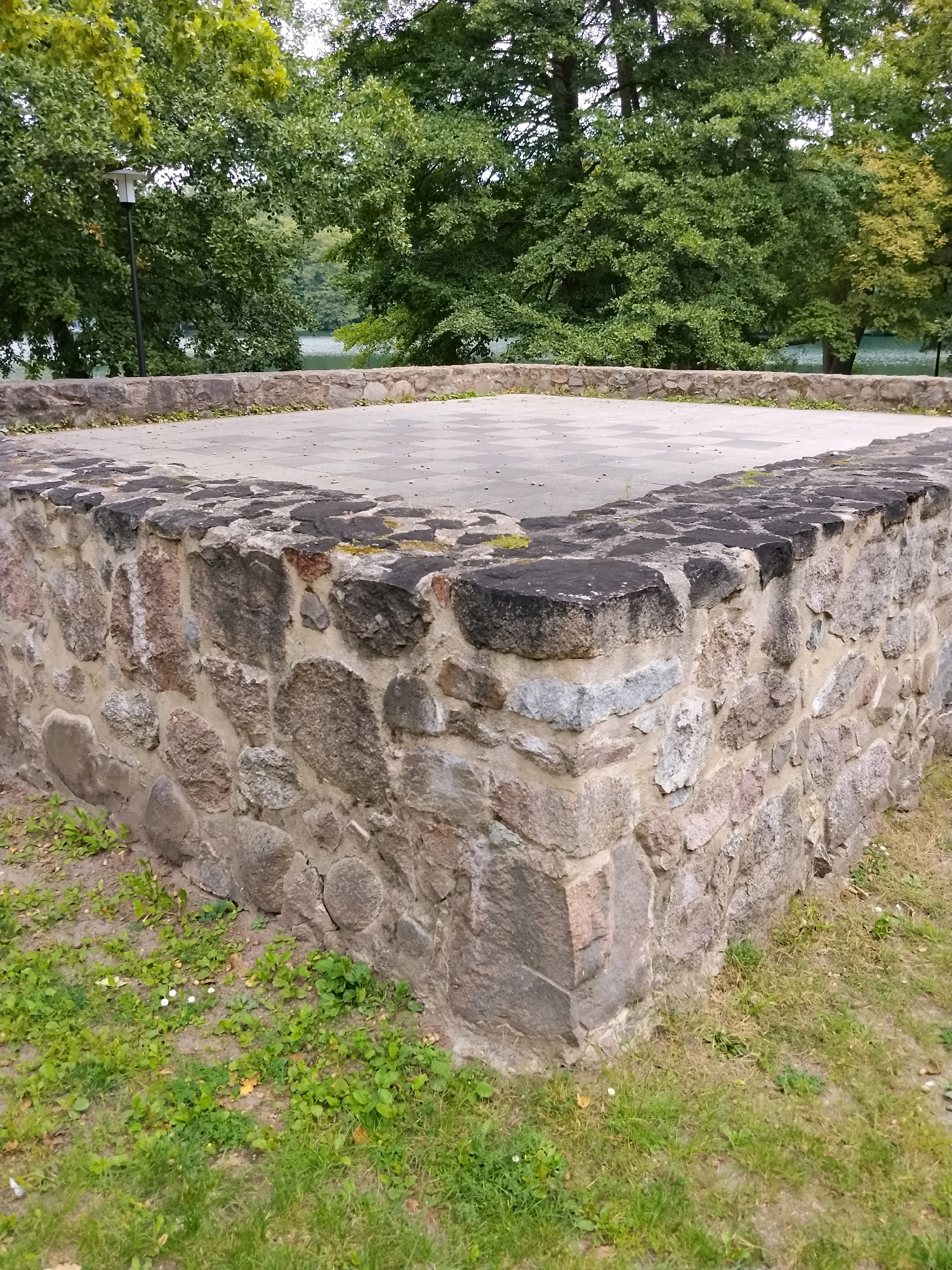 The image shows the partially ruined stone foundation of a structure in Łagów, Poland. The foundation is made of rough-hewn stones of various sizes and colors, mortared together. The top of the foundation is flat and paved with light-colored square stones, forming a raised platform. Green grass and some small white flowers grow around the base of the stone structure. In the background, lush green trees are visible, with a body of water and a cloudy sky beyond. A single modern lamppost stands to the left of the foundation, partially obscured by trees. The overall scene depicts an outdoor, historical site.