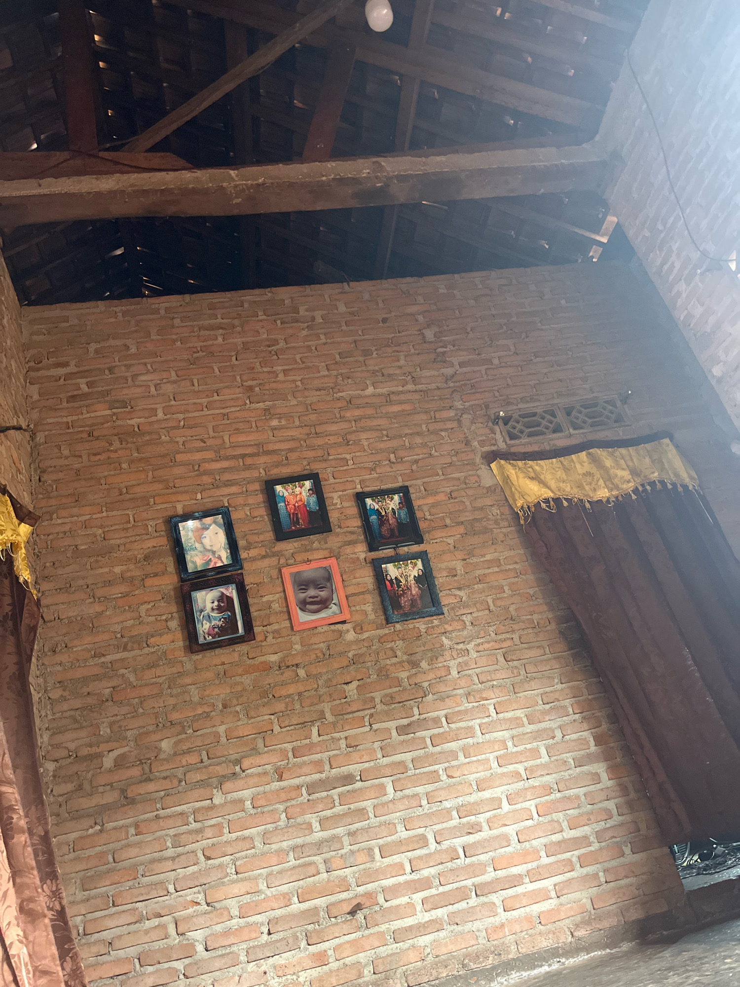The image shows the interior of a room with a brick wall and a wooden roof structure. There are several framed photographs hanging on the brick wall. The photographs display images of people, including children and families. Two dark brown curtains with a yellow valance are visible on the right side of the image, partially obscuring the wall and a ventilation grate above them. On the left side of the image, a portion of a brown curtain is also visible. The floor is not clearly visible, but appears to be a light gray concrete or stone. The overall impression is of a simple, possibly rustic, living space. The image is taken from a low angle, looking upwards towards the roof. The location is identified as Sidomulyo, Indonesia.