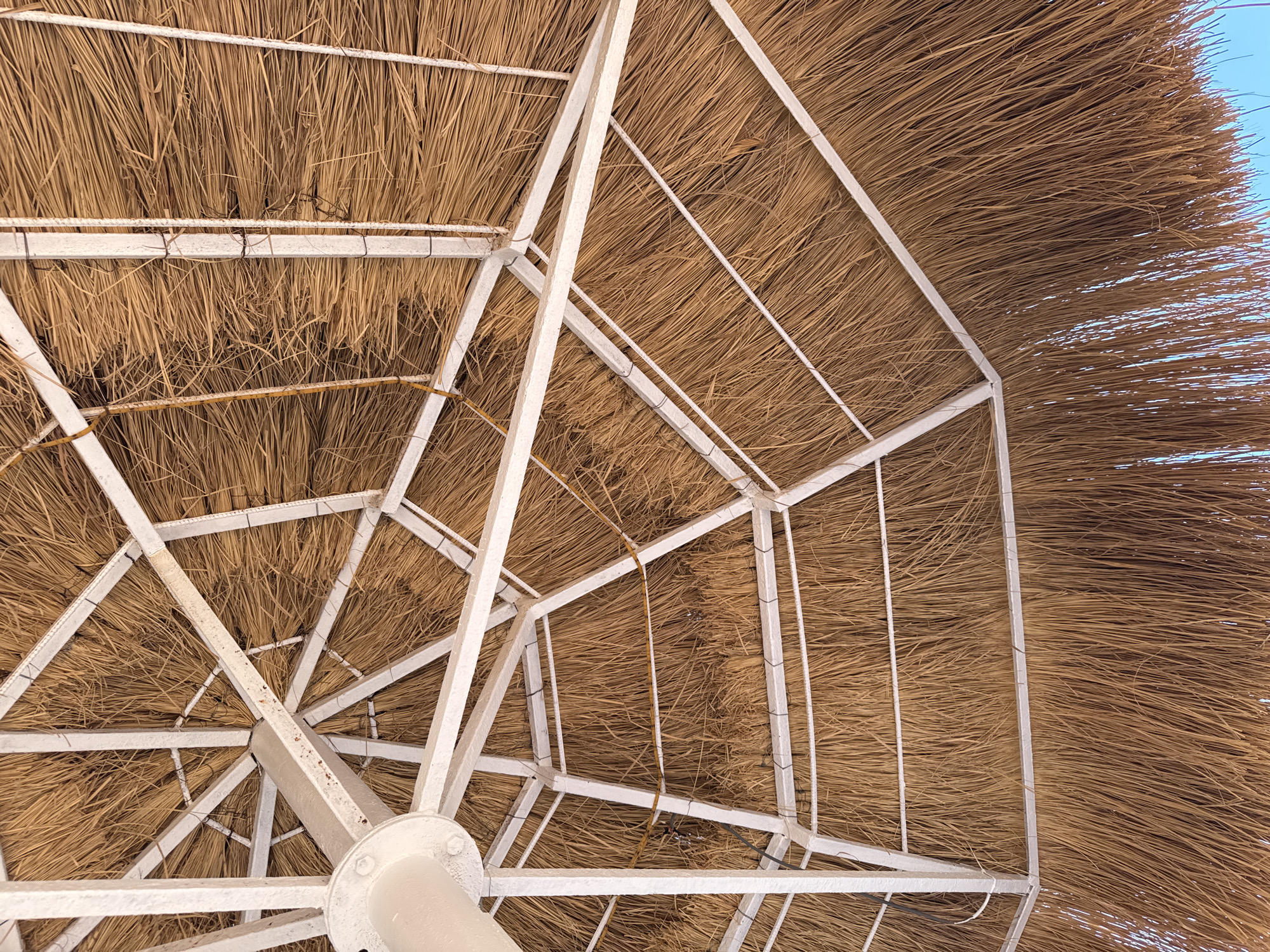 An underside view of a straw umbrella reveals a white metal frame supporting a thatched roof. The frame is composed of intersecting beams and struts, forming a polygonal structure. The straw, a dry, golden-brown material, is densely packed and hangs down in strands, creating a textured canopy. Small wires are visible, securing the straw to the frame. The background shows a clear, bright blue sky, suggesting an outdoor setting, likely in a warm climate. The image is captured from a low angle, looking upwards into the umbrella. The overall impression is one of rustic construction and simple design. The location is unknown, but it is likely a beach or a resort area in Egypt, given the style of the umbrella.