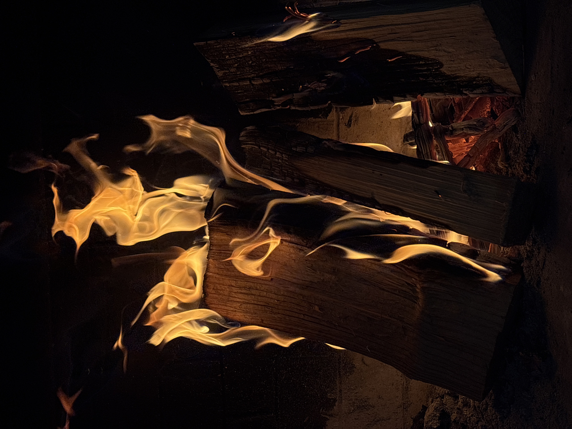 A fire burning in a fireplace. The fire is built of logs. The flames are yellow and orange. The background is dark.