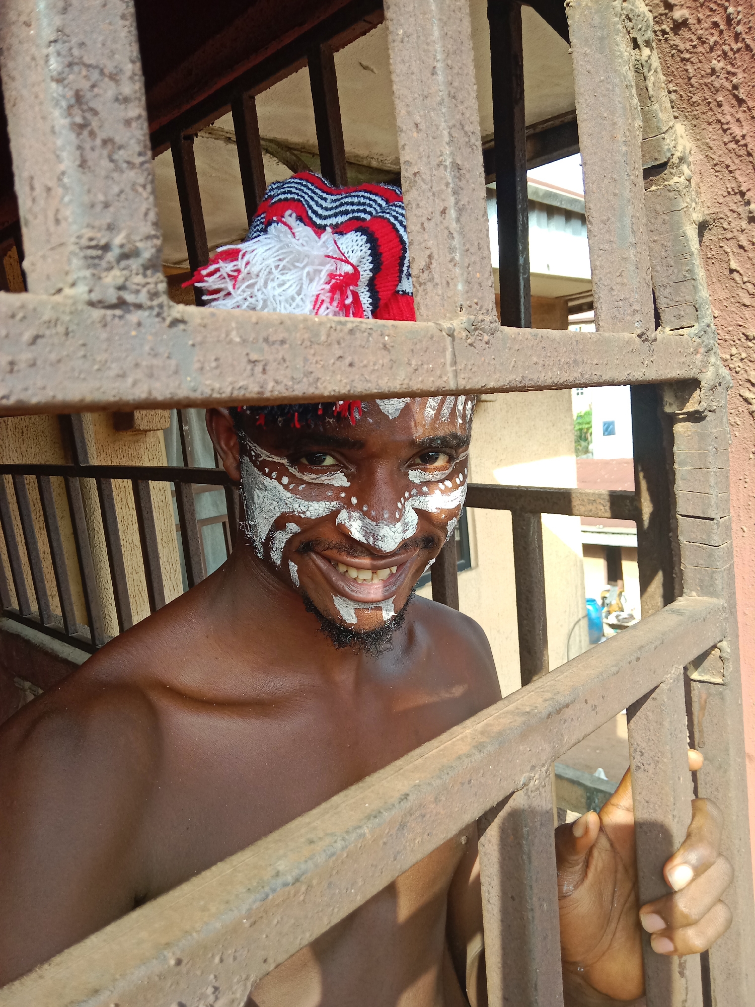 A medium, eye-level shot shows a bare-chested, dark-skinned man with white face paint, smiling through a rusty, metal security gate. The man wears a red, black, and white knit hat with white tassels. He grips the gate with his right hand. Beyond the gate, an exterior view suggests a residential area, possibly in Nkpor, Nigeria. A building with brown corrugated roofing is seen, along with another building painted in light yellow.