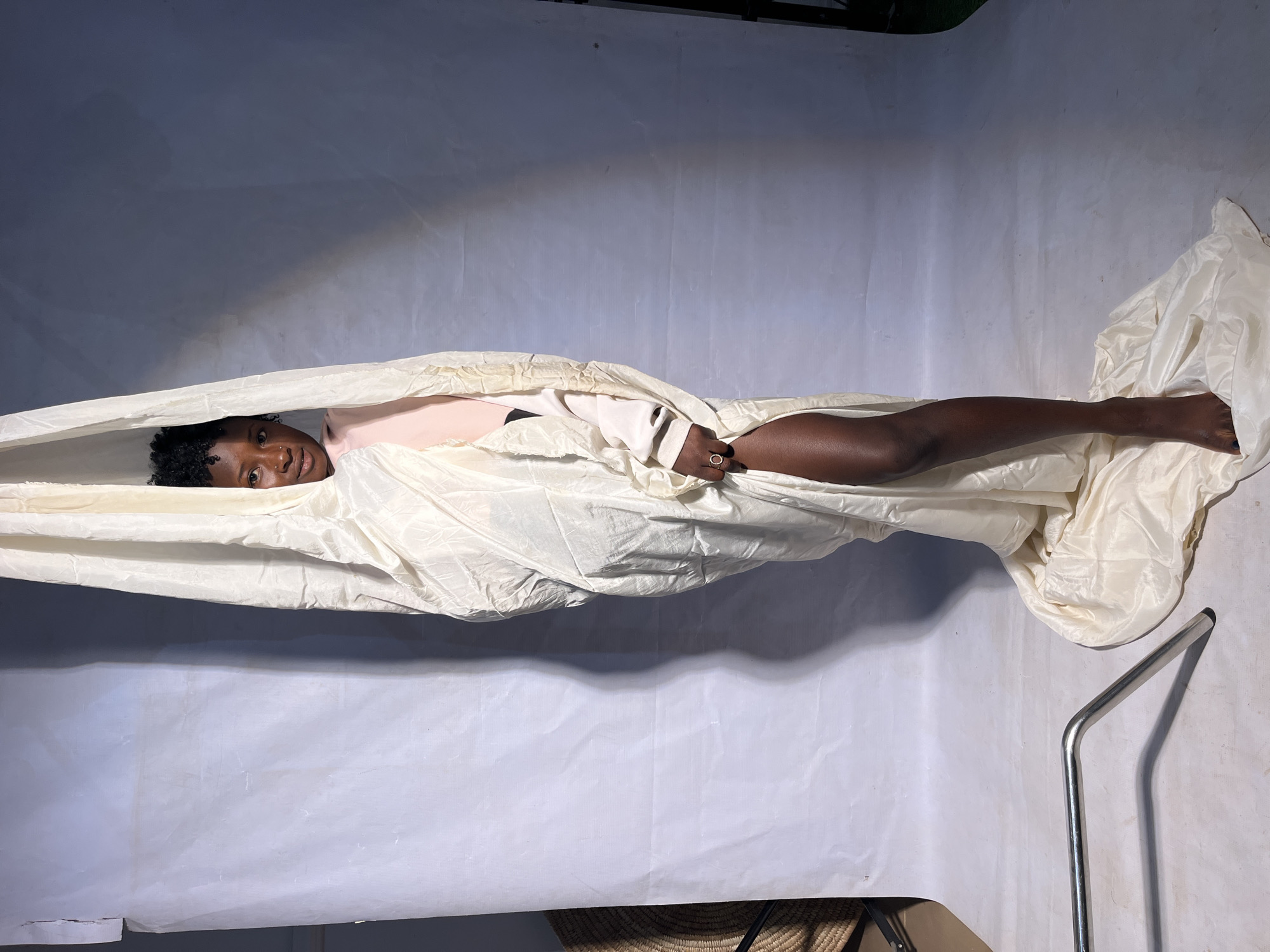 A young Black woman is wrapped in a large, cream-colored cloth, which drapes around her body and pools on the floor. She is standing, with one leg exposed and bent, revealing her knee and thigh. She is wearing a pink top and a ring on her left hand. Her hair is dark and curly. The background is a simple studio setting with a white backdrop and grey shadows. A silver metal stand is visible in the lower left corner. The lighting is soft and diffused. The location is Unknown, Unknown.