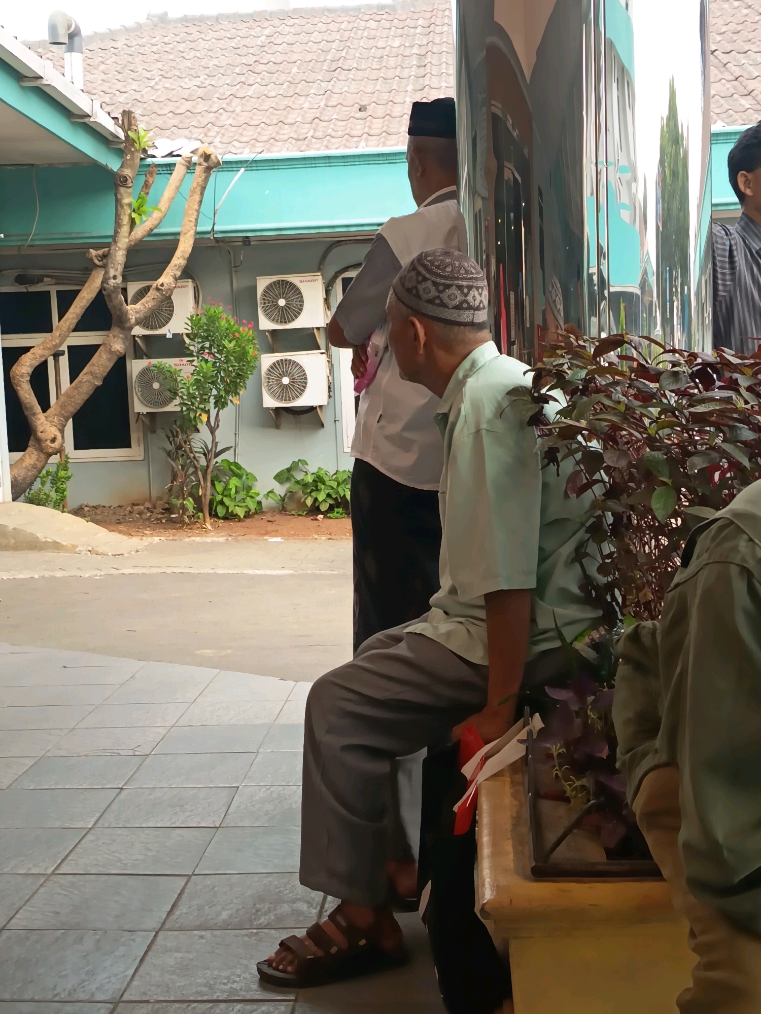 The image depicts a scene outside a building, possibly a mosque or public institution, in Tangerang, Indonesia. Three men are visible, two standing and one seated. The seated man, in the foreground and facing left, wears a light green short-sleeved shirt, grey trousers, and brown sandals. He holds a red and white object in his right hand. A man standing behind him and to his left, also facing left, wears a white shirt and a black peci (traditional cap). Another man stands further back and to the right, partially obscured, wearing a striped shirt. The environment includes an outdoor tiled area, a building with teal trim and tiled roofing, several outdoor air conditioning units mounted on the wall, a tree with exposed branches, and decorative plants. A reflective pillar is also present, showing distorted reflections of the surroundings. The overall impression is that of people waiting outdoors.