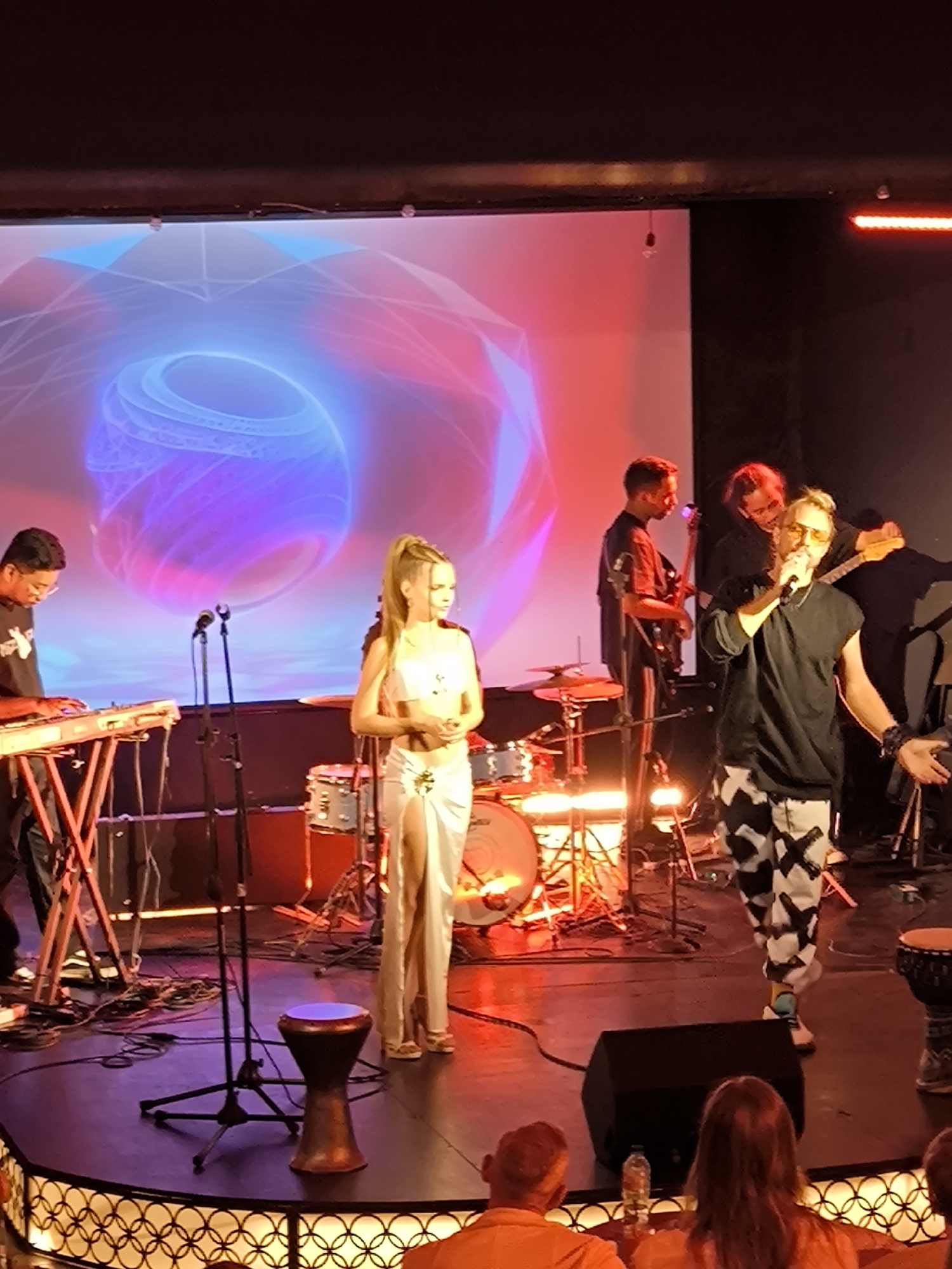 The image shows a live musical performance on a stage, likely indoors. A female vocalist in a light-colored dress stands near the center, while a male vocalist in a black shirt and patterned pants is positioned to her right. Several other musicians are present, playing instruments such as keyboard, bass guitar, and drums. A large screen behind the band displays abstract graphics with blue and pink colors. The stage is illuminated with stage lighting. In the foreground, the backs of the heads of audience members are visible. The location is unspecified, although the prompt mentions Kediri, Indonesia.