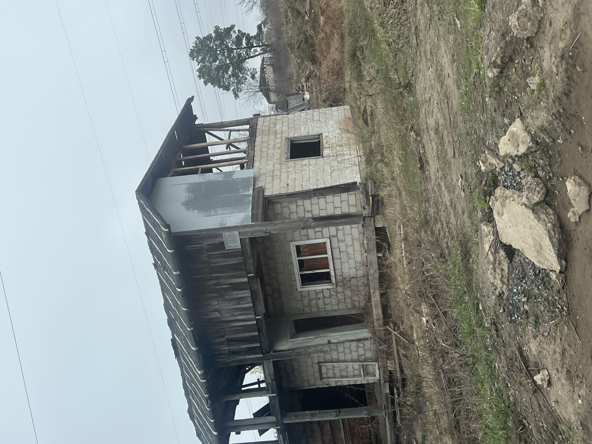 The image, taken in Chaikovsky, Russia, captures a two-story, unfinished or derelict building under an overcast sky. The building's walls are constructed from light-colored cinder blocks, with visible wooden structural elements forming the roof frame and supporting the upper floor. Several window openings are present; some are empty, while others contain partial frames or glass. The overall structure appears to be in a state of disrepair or halted construction, with exposed wooden beams and partially covered sections.
In the background, overhead power lines stretch across the grey sky. A large, mature pine tree stands prominently to the right of the power lines, beyond a distant hill covered in sparse, dry vegetation. The ground surrounding the building is uneven, composed of mud and dirt with patches of dry grass and some green shoots. To the immediate right of the structure, there is a pile of concrete debris and scattered gravel.
The scene conveys a sense of abandonment or neglect. The weather appears to be overcast during daytime, with no strong shadows cast. There are no people visible, and no specific activities are taking place. No discernible text is present in the image. The original image is heavily tilted to the right.