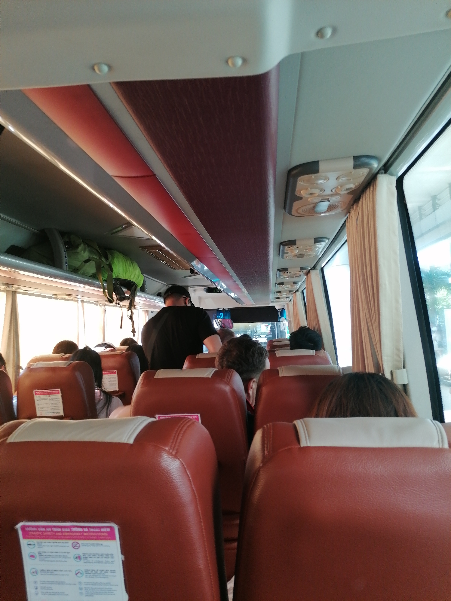 The image depicts the interior of a passenger bus in Bac Cam Ranh Ward, Vietnam. The bus is mostly full, with passengers seated in brown leather seats with white headrests. Some seats have safety instruction cards attached. An individual in a black shirt is standing near the front, possibly moving towards the front or rear of the bus. Luggage, including a green backpack, is visible in the overhead storage compartment. Curtains are present by the window.