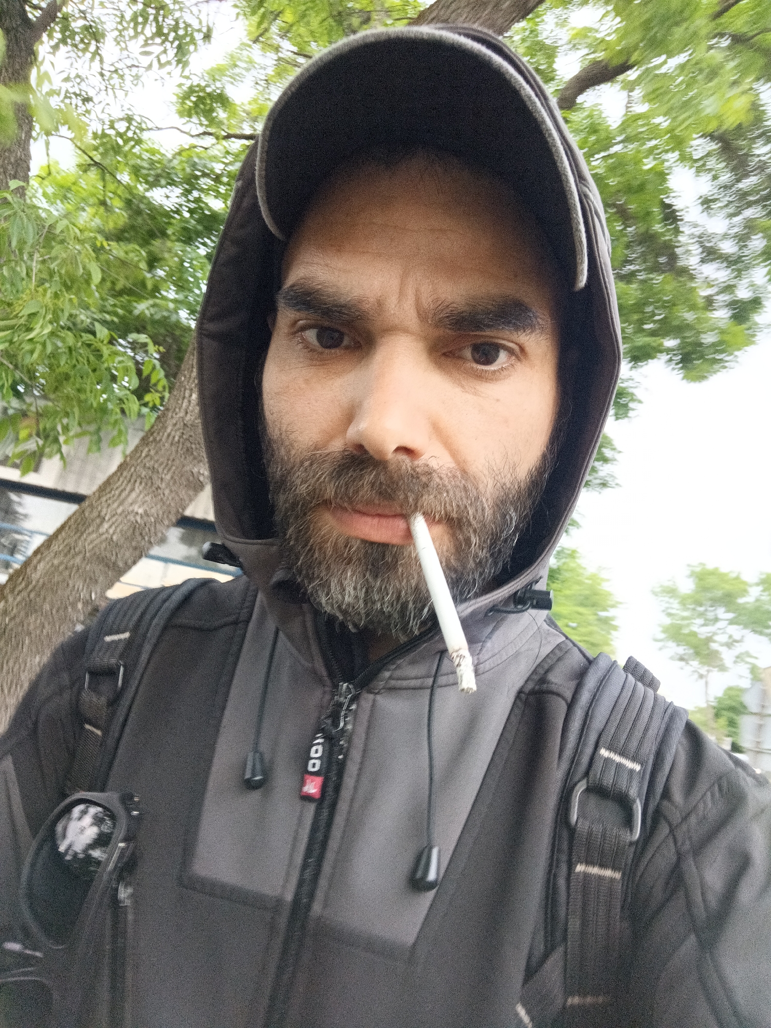 A man stands outside in the city of Unknown, Unknown. He has a beard and wears a dark grey hooded jacket with a zipper. The hood is up, and a dark baseball cap sits underneath. A cigarette is held in his mouth. He wears a backpack. A tree and green foliage are visible in the background.