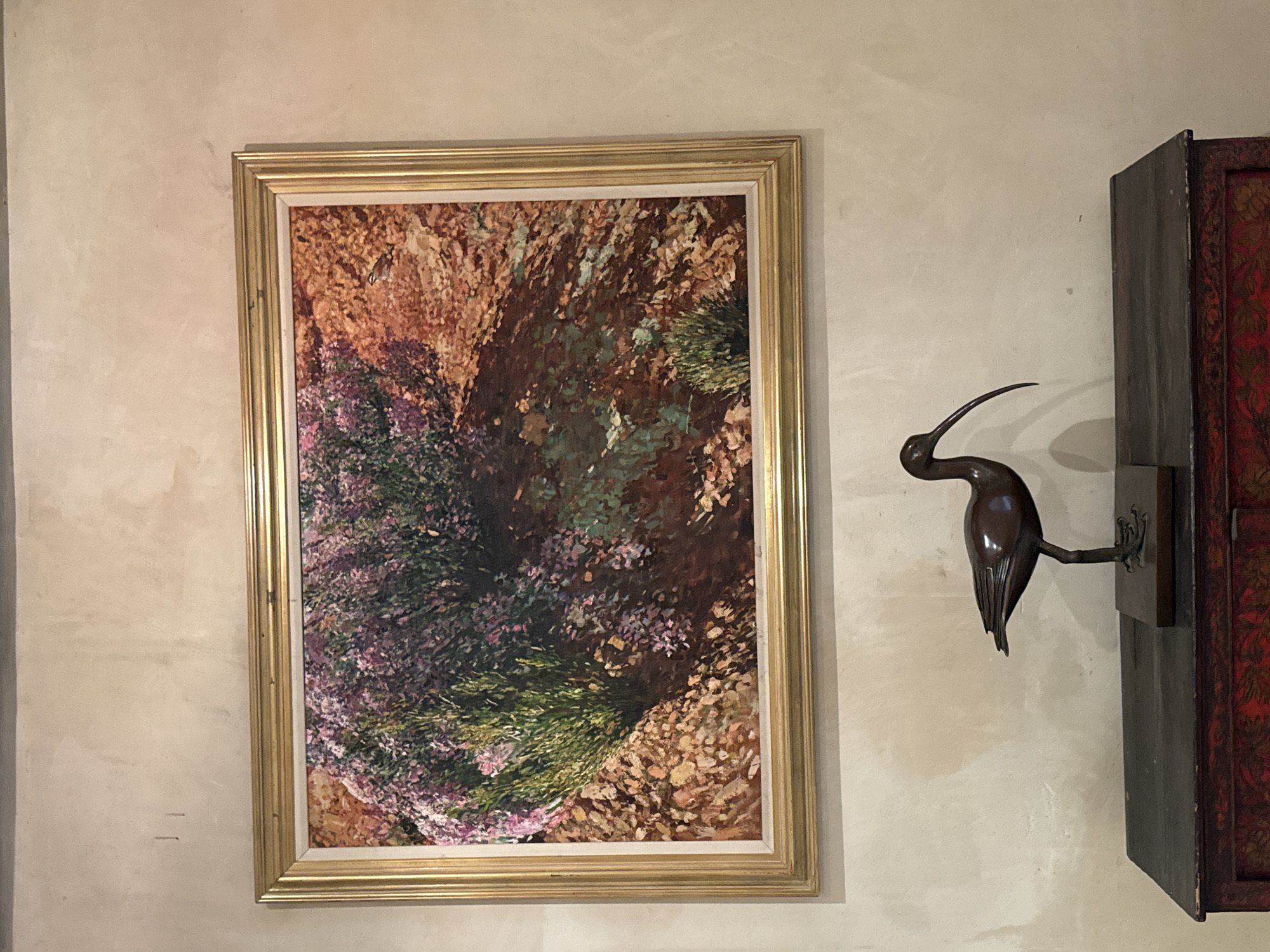 A medium shot of a framed painting hanging on a light beige wall, next to a dark brown decorative object resembling a bird. The painting is in a gold frame and depicts a landscape of brown, green, and purple hues. The wall has a textured appearance. The bird decoration is mounted on a dark, rectangular block next to a cabinet with a reddish, patterned door. The location is indoors. There are no people present. There is no indication of this being in Stellenbosch Nu, South Africa.