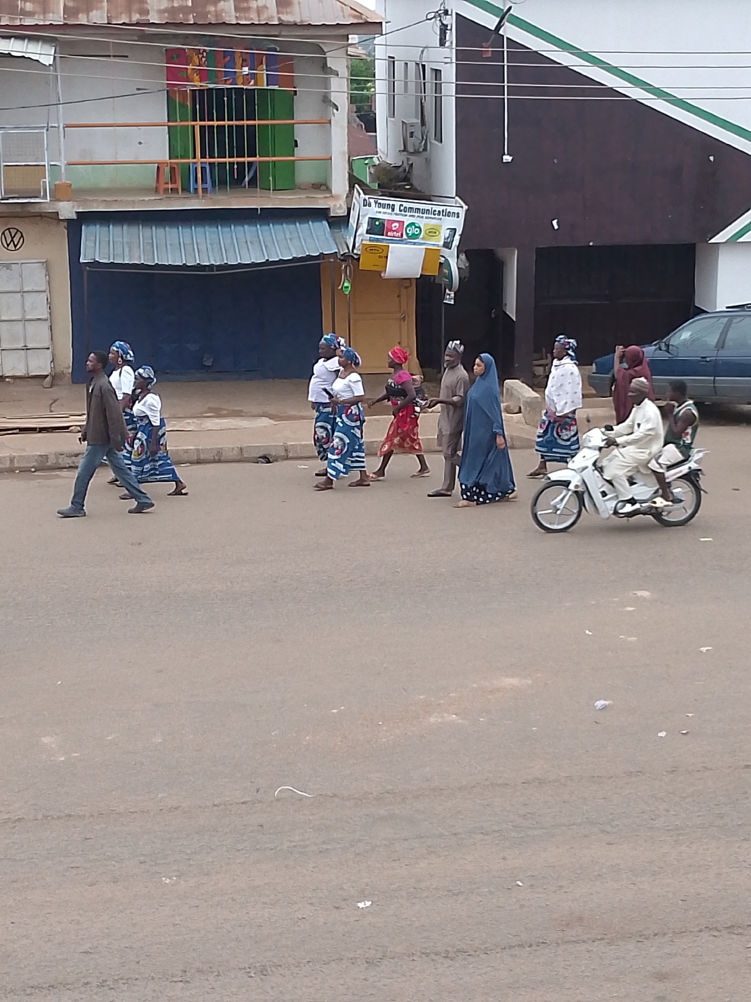 The image depicts a street scene in Bukuru, Nigeria. A group of people are walking along the road, with a man on a motorcycle carrying a passenger approaching from the right. The pedestrians are dressed in a variety of traditional and contemporary clothing. Buildings line the street, including a two-story structure with a blue awning and a sign for "Da Young Communications". A car is parked near the building on the right side of the frame. The road surface appears to be paved and worn.