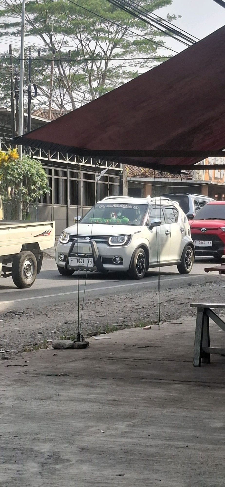 A white Suzuki Ignis is parked on the side of a street in Rancagoong, Indonesia, with a red car and a white utility vehicle also visible. The white car has a black bull bar attached to its front bumper, and a license plate that reads "F 1041 YH". Inside the car, a person wearing a green shirt is visible in the driver's seat. The street is paved with asphalt and has some debris on it. In the background, there is a large metal gate, trees, and utility poles with wires. The scene is partially covered by a large, dark red tarp.