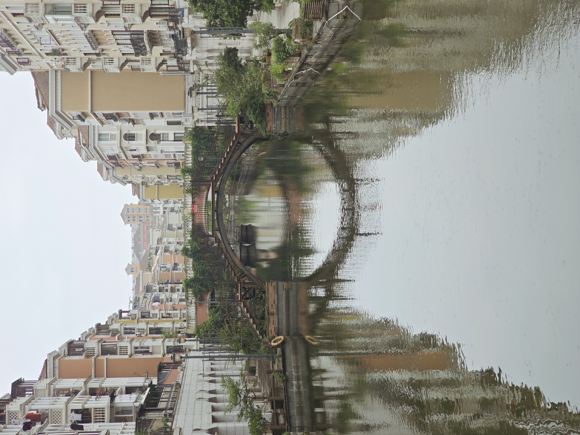 The image depicts a canal lined with colorful buildings, characteristic of the European-style architecture found in the Venetian Palace residential community in Xiaoshan District, China. The canal itself reflects the buildings and a decorative arched bridge that spans across it. The bridge has a rustic metal railing and wooden steps leading up to it, with lush greenery growing along its sides. On the canal banks, there are lampposts, potted plants, and benches. The buildings are multi-story, with balconies and terracotta-tiled roofs, painted in shades of orange, yellow, and cream. The sky is overcast, casting a soft, diffused light on the scene. The water in the canal is calm, with ripples creating distorted reflections of the surrounding structures. There are no people visible in the image.