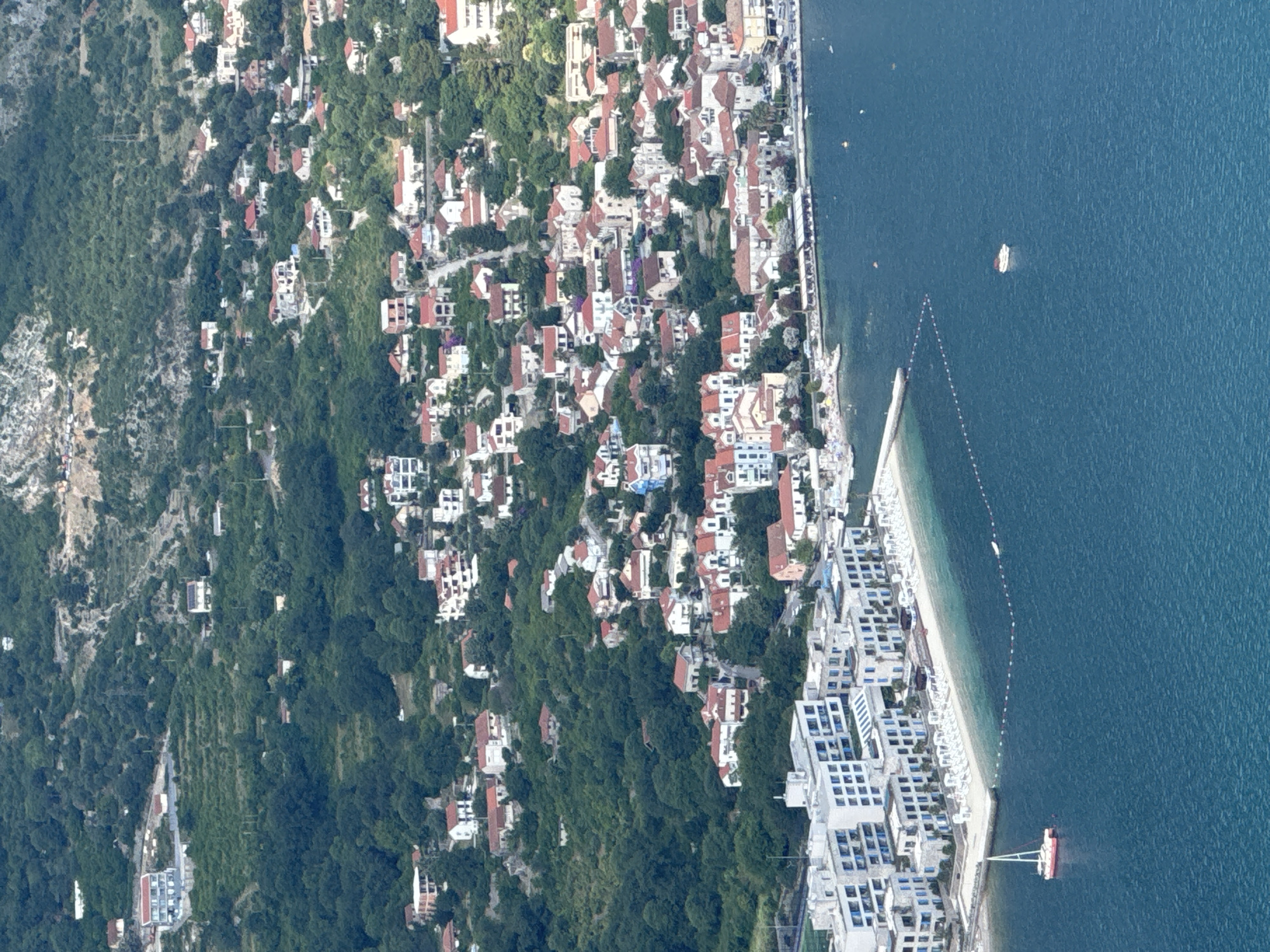 The aerial image depicts a coastal scene in Strp, Montenegro, showcasing a cluster of buildings nestled on a hillside overlooking a bay. In the foreground, a modern resort complex with white facades and blue-tinted windows stretches along a sandy beach, dotted with numerous white lounge chairs and umbrellas. A red and white sailboat with its sail raised is anchored in the clear blue water near the shore. Further out in the bay, a white boat is visible. The hillside behind the resort is densely populated with residential buildings, characterized by red-tiled roofs and white or light-colored walls. These houses are interspersed with lush green vegetation. The terrain rises steeply, with some buildings appearing to be constructed on terraces. The overall impression is one of a well-developed coastal town with a mix of modern and traditional architecture, situated in a scenic, hilly landscape.