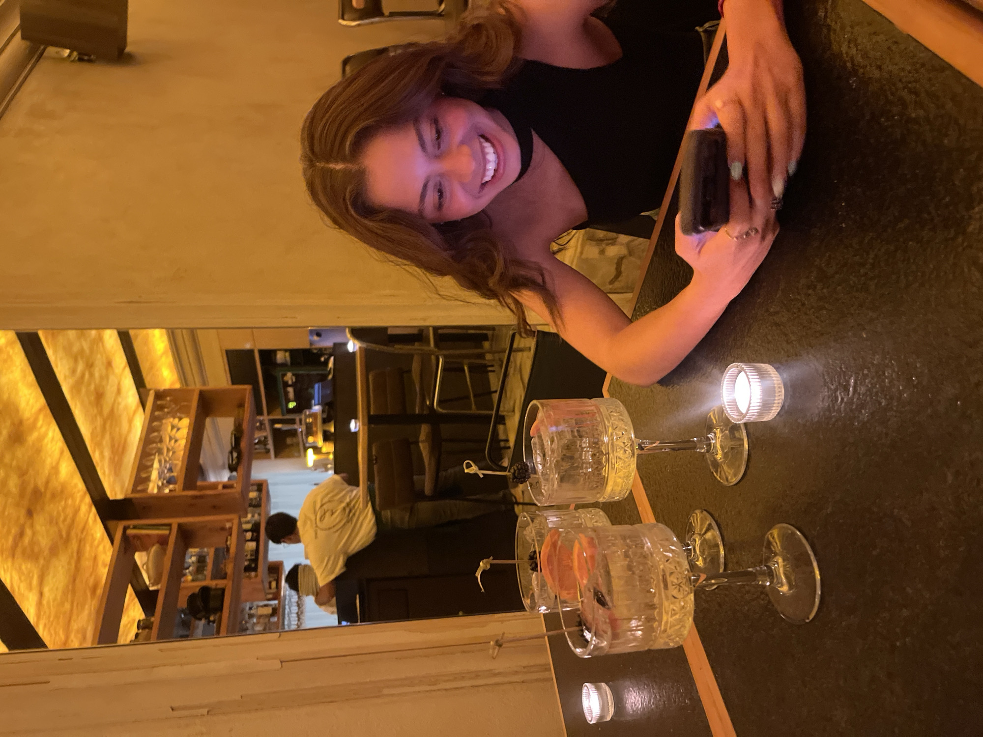 A woman in a black top smiles and holds a black smartphone at a bar with two cocktails and a lit tealight candle. Shelves with glassware are visible in the background. A man in a white shirt stands near the back of the bar. This image was likely taken in San Andrés Cholula, Mexico.