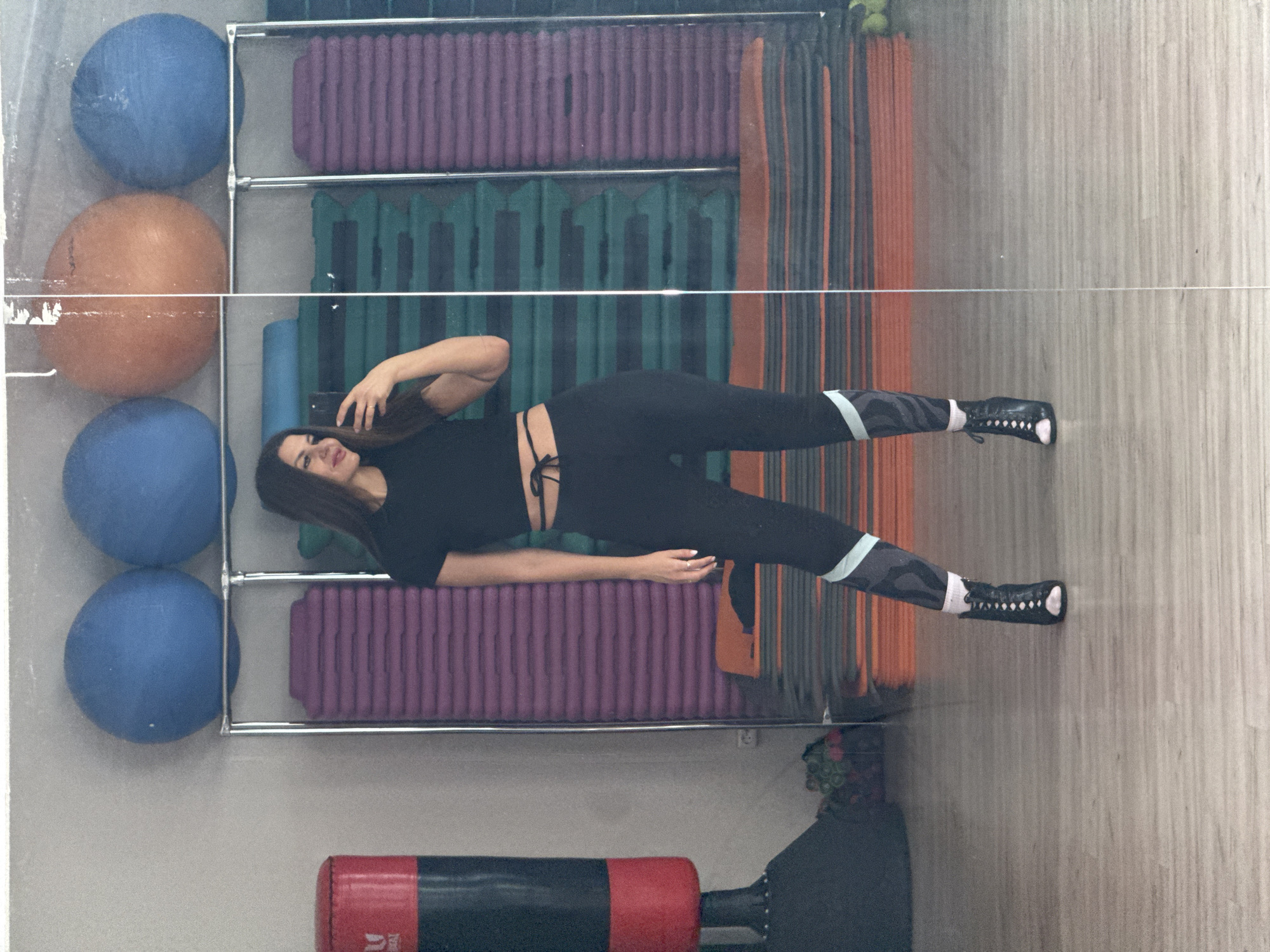 The image shows a woman lying on her side in front of a mirrored wall in a gym in Chaikovsky, Russia. She is wearing a black top, black leggings with light blue accents, and black shoes with white socks. The reflection shows gym equipment including exercise balls, step platforms, and a punching bag. She appears to be holding a phone and taking a picture of herself in the mirror.