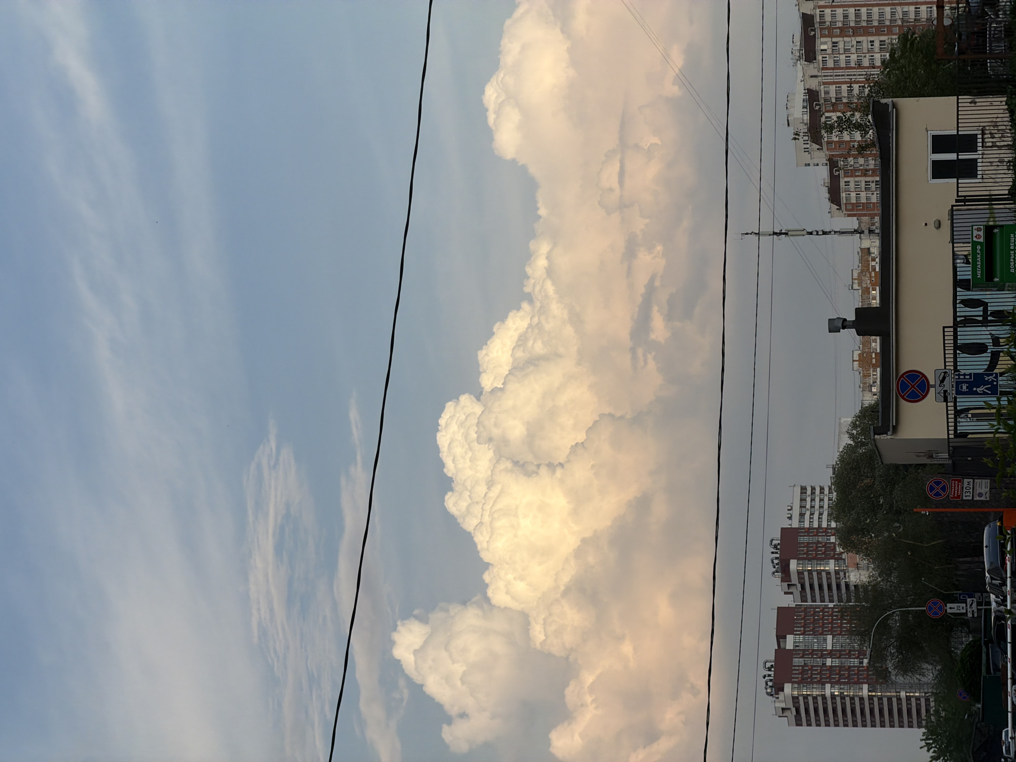 The image shows an urban scene, likely in Moscow, Russia, judging by the architectural style and signage. The sky is partly cloudy with a large, billowing cumulus cloud dominating the view. Utility lines run across the sky. Buildings are visible in the lower portion of the image, including high-rise residential buildings and a smaller, light-colored building with signage in Russian. Street signs are also visible, including "no parking" signs. The buildings suggest a residential area. The orientation of the image is vertical.