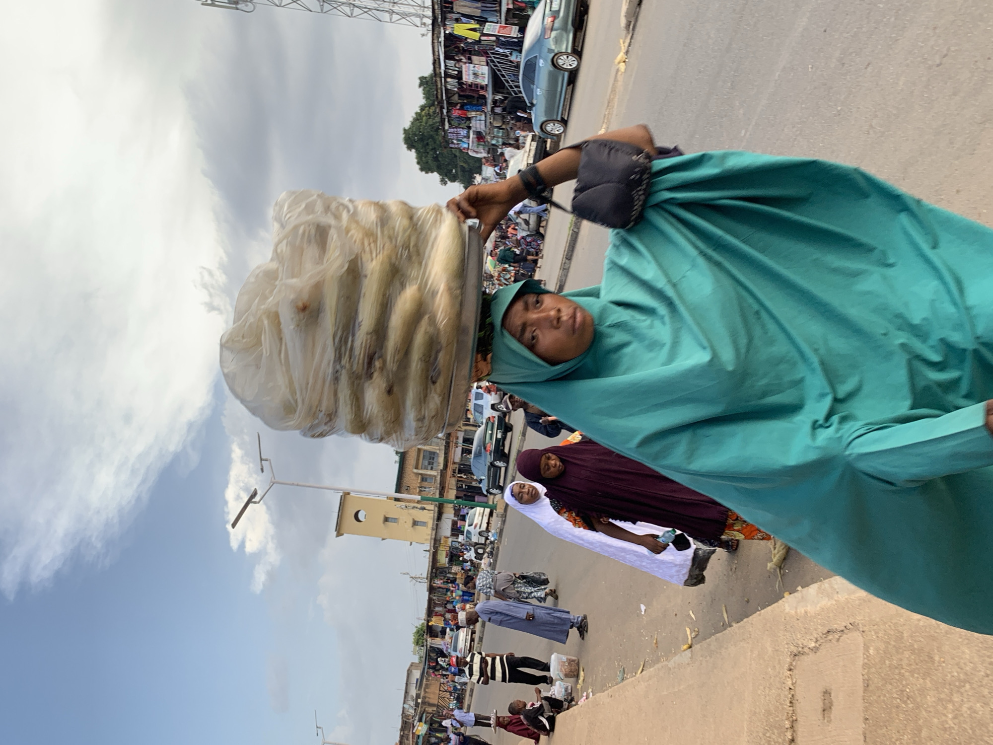 In Jos, Nigeria, a woman dressed in a turquoise hijab carries a large bundle of goods, possibly produce, on her head. The bundle is wrapped in clear plastic and rests on a metal tray. She is walking along a dusty, paved street lined with market stalls and shops. Other people are visible in the background, some also carrying items and walking, while others are seated near the roadside. Cars are present on the street, and a telecommunications tower can be seen in the distance. The sky is partly cloudy. The scene appears to be a busy marketplace on a typical day.