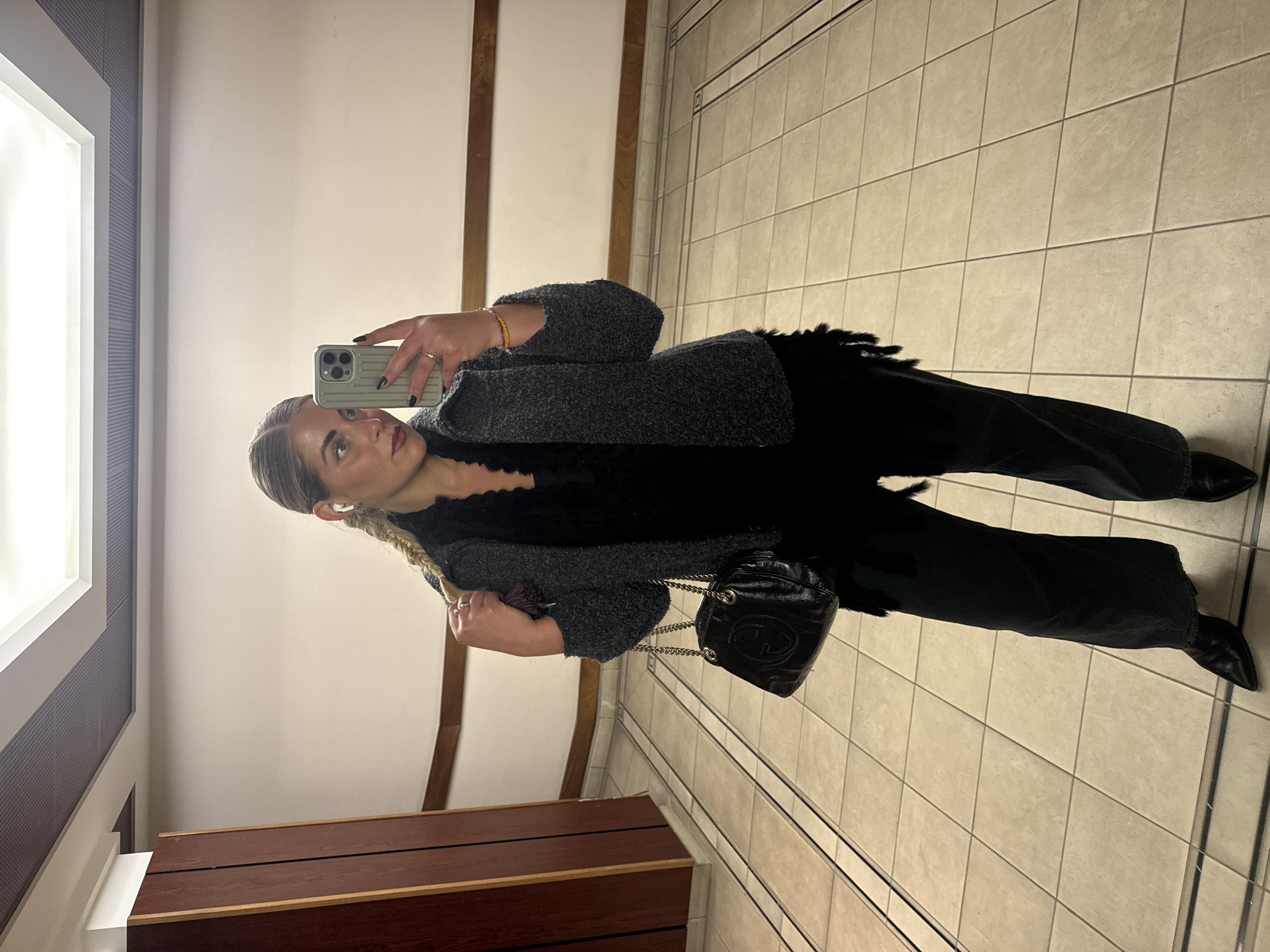 A woman is taking a selfie in a mirror. She is wearing a gray, tweed jacket, a black top with fringed detailing, black jeans, and black pointed-toe boots. She has a black leather purse with a gold chain strap. Her hair is blonde and styled in a braid. The background suggests she is in a restroom with tiled walls and a wooden vanity. The setting could be in Paris, France, though there are no explicit landmarks to confirm it.