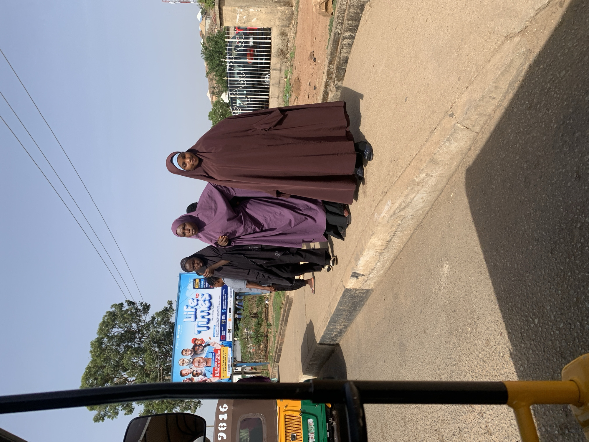 The image shows a street scene in Jos, Nigeria. Three women are walking on the sidewalk, wearing traditional Muslim attire: the first woman wears a brown hijab, the second a purple hijab, and the third a black hijab. To the left of the women is a billboard advertising a theatrical event titled "Life Times", featuring pictures of people. A tree is visible behind the billboard. Overhead power lines are strung along the street. The view is from inside a vehicle, specifically a yellow and green tricycle taxi, with its front frame visible in the foreground. The street surface is paved and has a distinct curb. A building with a metal gate is situated behind the women.