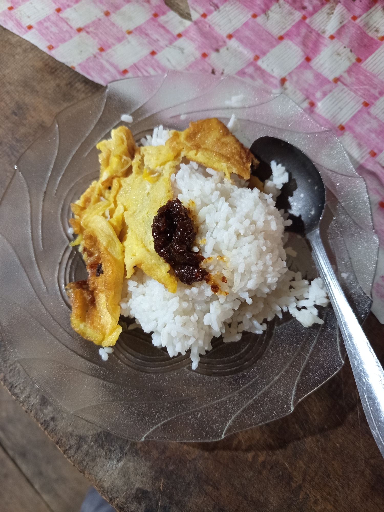An overhead shot shows a portion of a meal on a clear, decorative plate, placed on a wooden table. The meal consists of white rice, topped with a dollop of dark, reddish-brown sambal or chili paste, and several pieces of fried egg. A dark-colored spoon rests on the right side of the plate. The background features a pink and white checkered fabric, possibly a tablecloth or placemat. The overall impression is of a simple, home-style meal. There is no indication of the specific location within the city of Unknown, Indonesia.