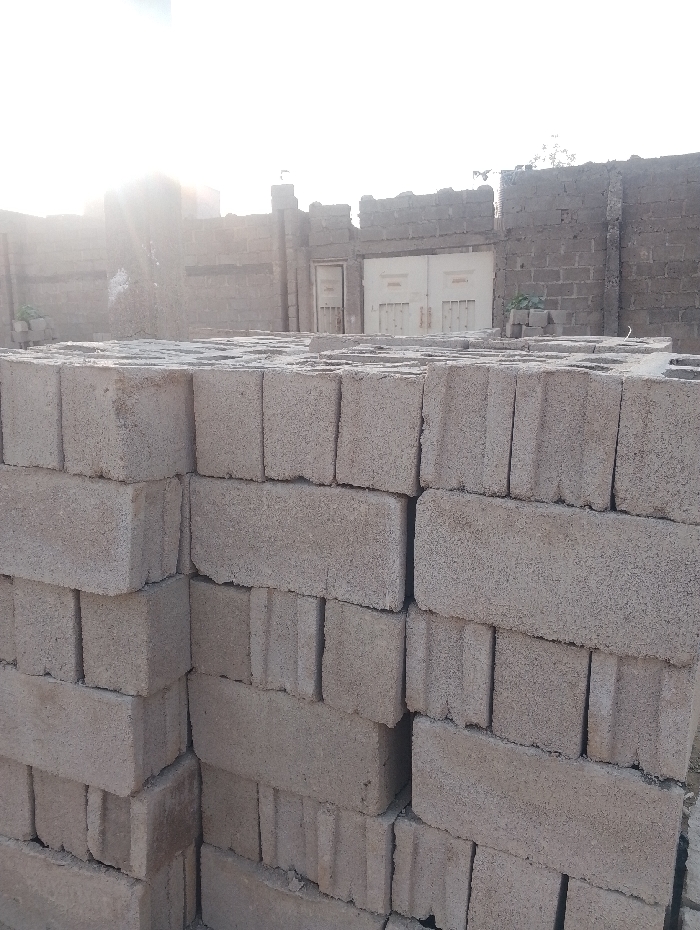 Stacks of gray concrete blocks fill the foreground. Each block has two vertical rectangular holes. Behind the blocks, a weathered brick or cinder block wall surrounds a yard. A closed, double gate is visible within the wall, flanked by single doors. The sky is partly visible in the upper portion of the image, with a bright light source partially obscured by the wall, casting a hazy glow.
