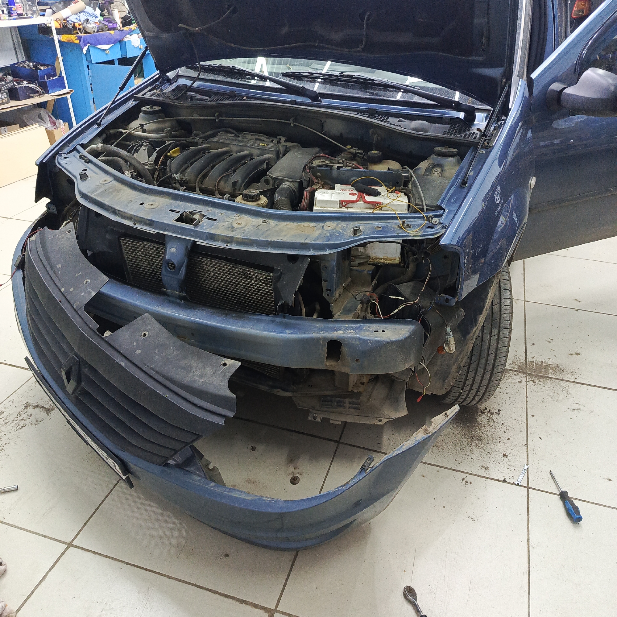 This image depicts the front end of a blue car, likely a Renault Dacia Logan, undergoing maintenance or repair within an indoor garage or workshop in Chaikovsky, Russia. The car's hood is open, held up by gas struts, revealing a dusty engine bay. Key engine components visible include a dark engine cover with "16V" embossed on it, various hoses and wiring, and a white AKOM brand battery with red terminals and yellow wires connected.
The car's front bumper is partially detached; its main body rests on the floor in front of the vehicle, exposing the radiator and other frontal structures behind it. The bumper itself and the visible inner workings of the car's front show significant dirt and grime.
The surrounding environment suggests a functional workspace. The floor is covered with light-colored tiles, marked with dirt, dust, and some dark stains. In the background, blue metal shelving units are visible, cluttered with assorted boxes, tools, and other workshop items. Several tools, including a blue-handled screwdriver, a ratchet, and a small wrench, are scattered on the floor around the car, indicating active work. The driver's side door is also open. No individuals are present in the image.